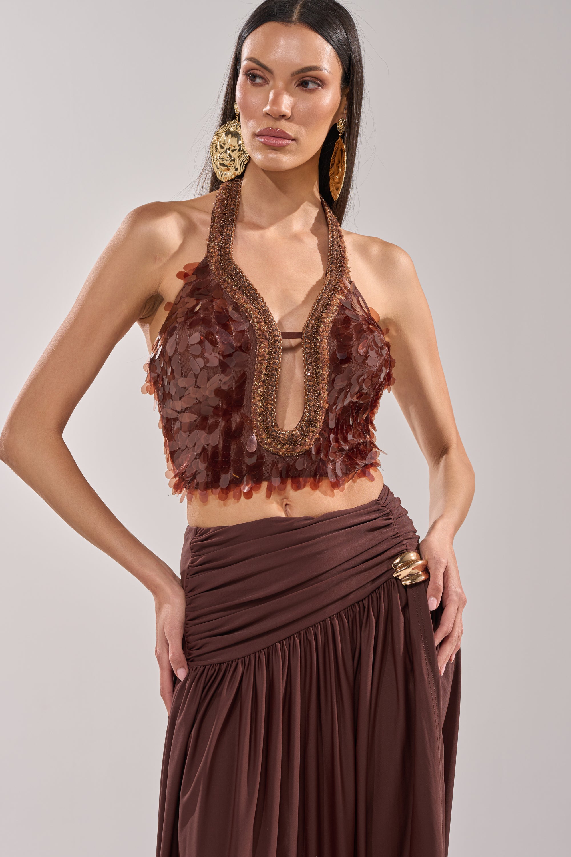 A woman poses against a plain background in the ISLAND GIRL SEQUIN BLOUSE, a sleeveless bronze top with textured embellishments and deep V-neck, paired with a high-waisted draped brown skirt and large gold earrings. 0
