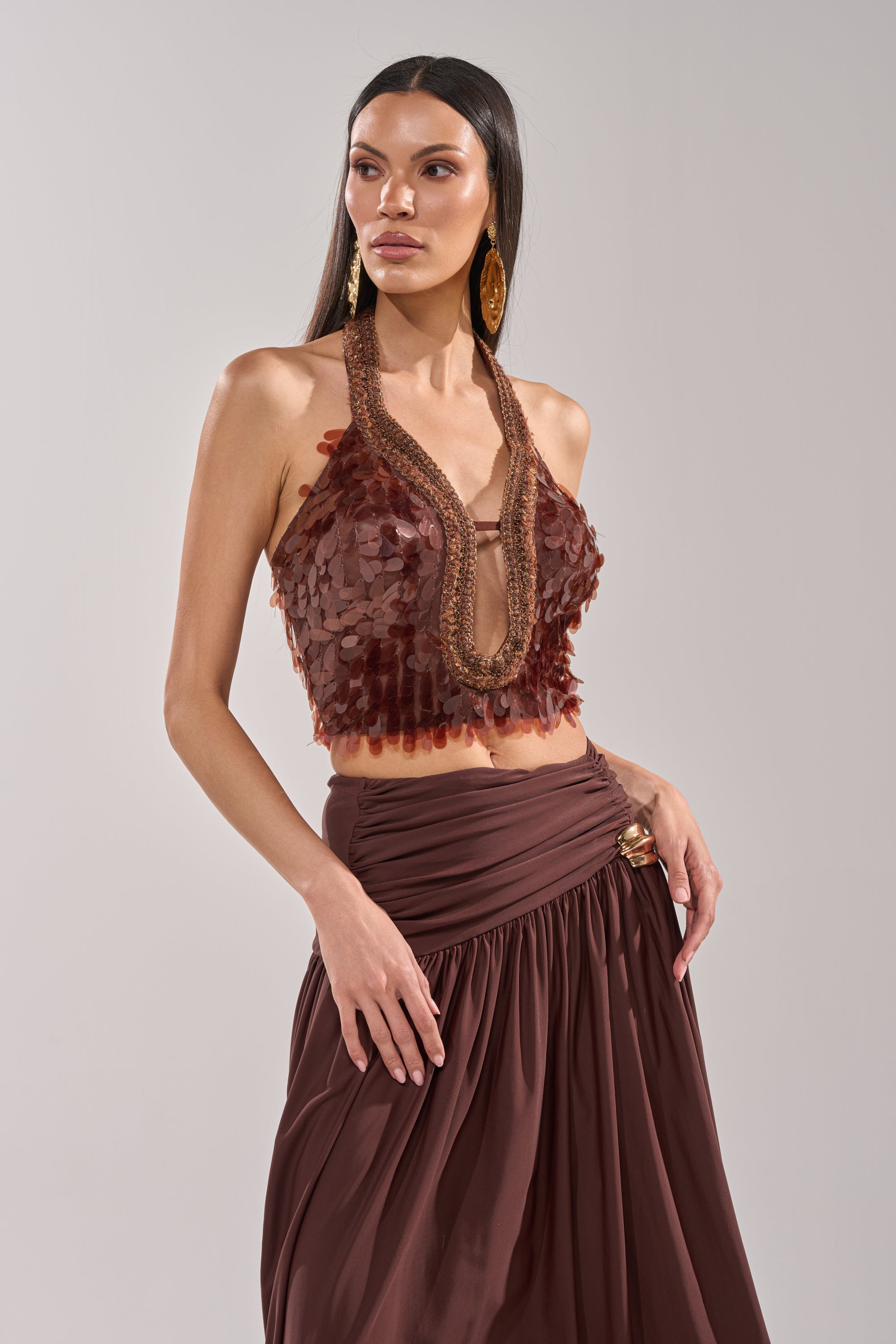 A woman poses against a plain background wearing the ISLAND GIRL SEQUIN BLOUSE, a bronze statement top with textured, petal-like details, paired with a flowing brown skirt and her hand on her hip. 1