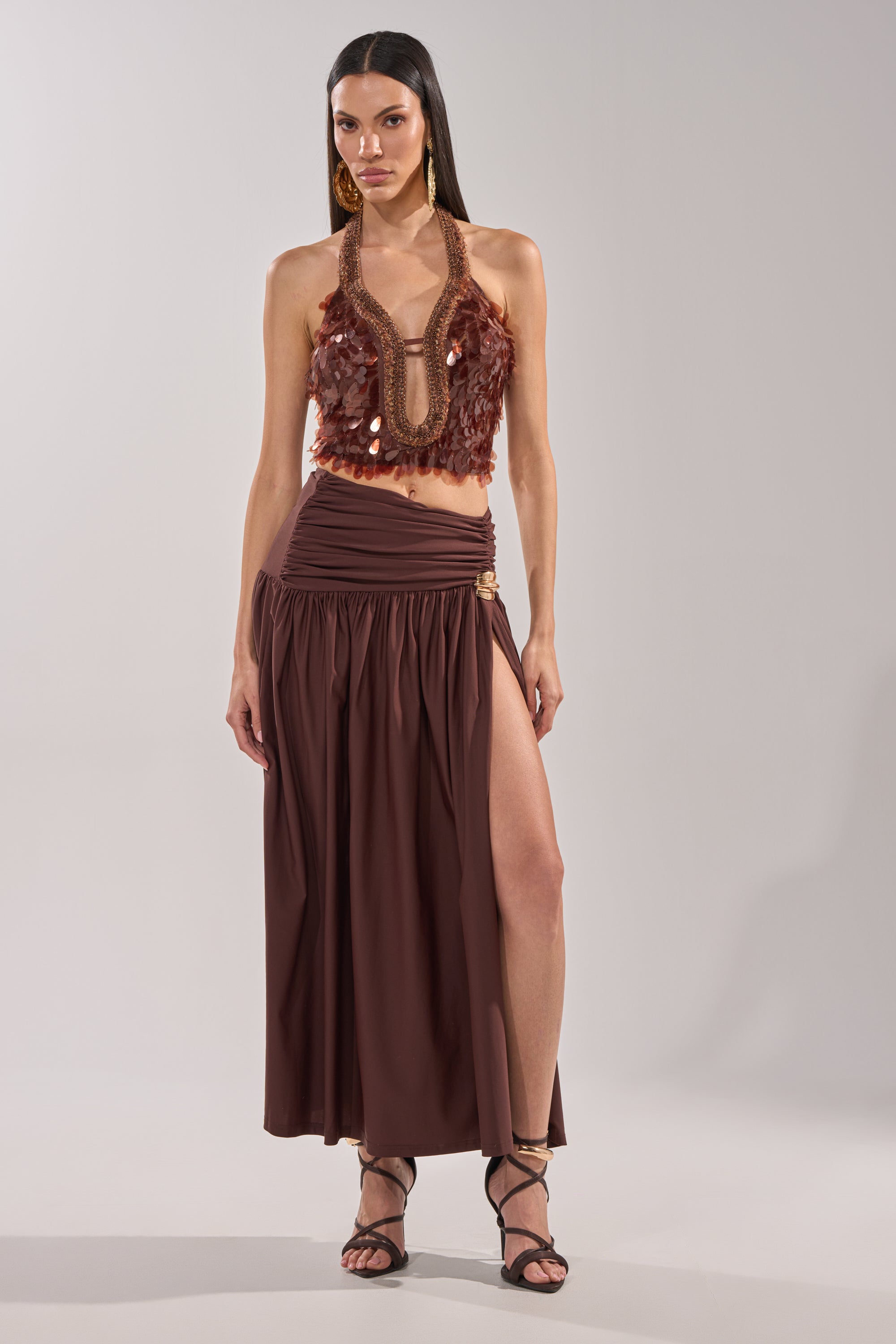 A woman with long dark hair and gold earrings poses against a light background, wearing the ISLAND GIRL SEQUIN BLOUSE—a bronze sequin halter top with fringe—paired with a matching high-slit maxi skirt and strappy heels. 4