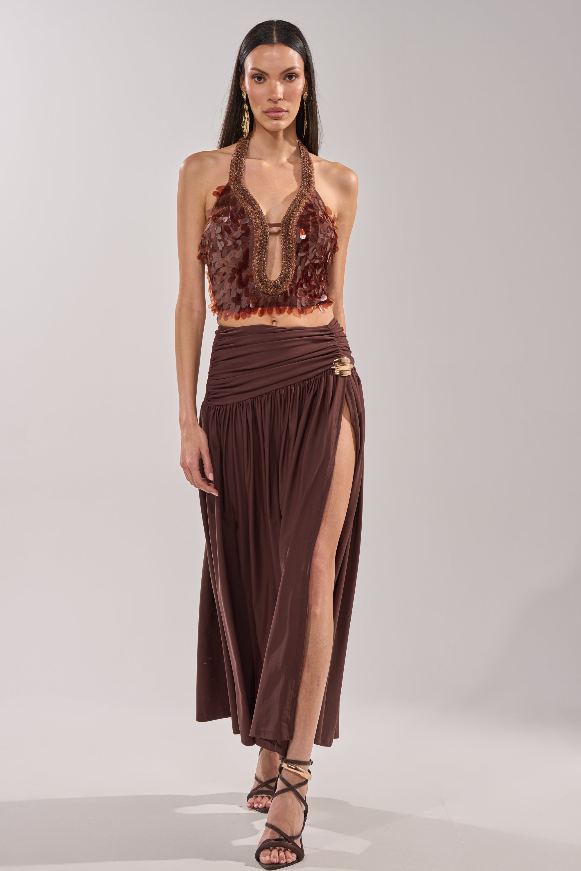 Wearing the ISLAND GIRL SEQUIN BLOUSE with textured bronze sequins and a matching high-slit skirt, a woman accessorizes with gold earrings and strappy sandals as she walks against a plain backdrop. 5