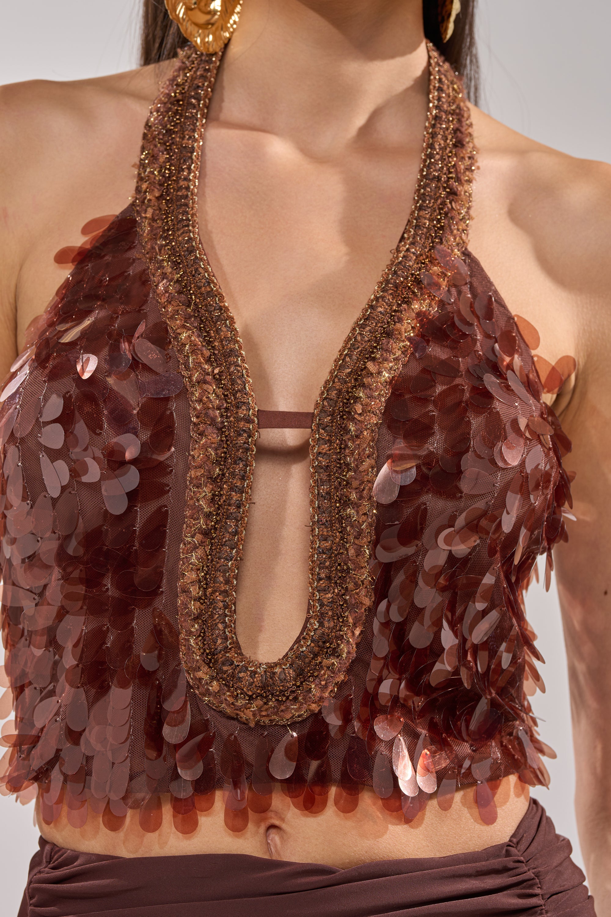 A woman wears the ISLAND GIRL SEQUIN BLOUSE, a party-ready bronze halter top with large shiny sequins, beaded trim, and a plunging neckline, paired with a matching brown skirt. Only her upper torso is visible. 6