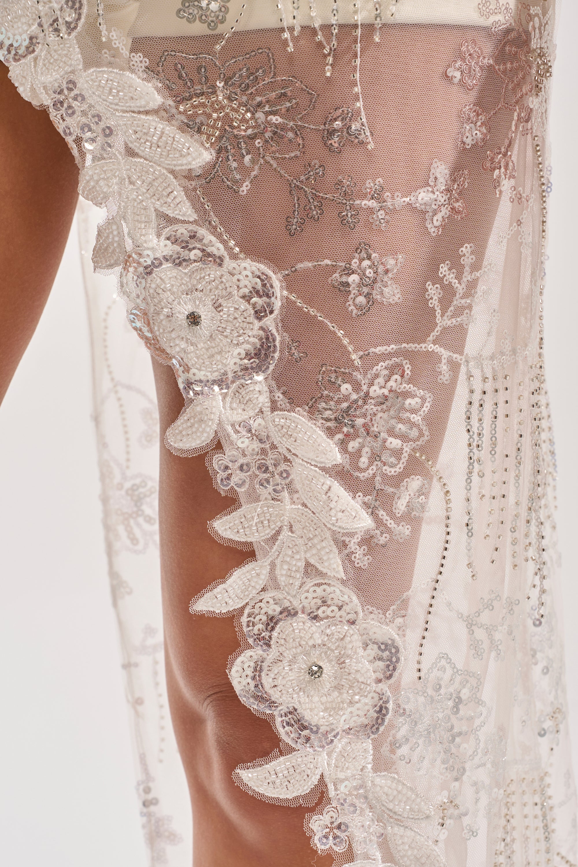 Close-up of sheer, embroidered lace with intricate floral and leaf motifs, sequins, and beads draped over a leg. Perfect for bridal evening wear or as the SHINE BRIGHT BLOUSE’s embellished asymmetrical top. Light plain background shown. 6