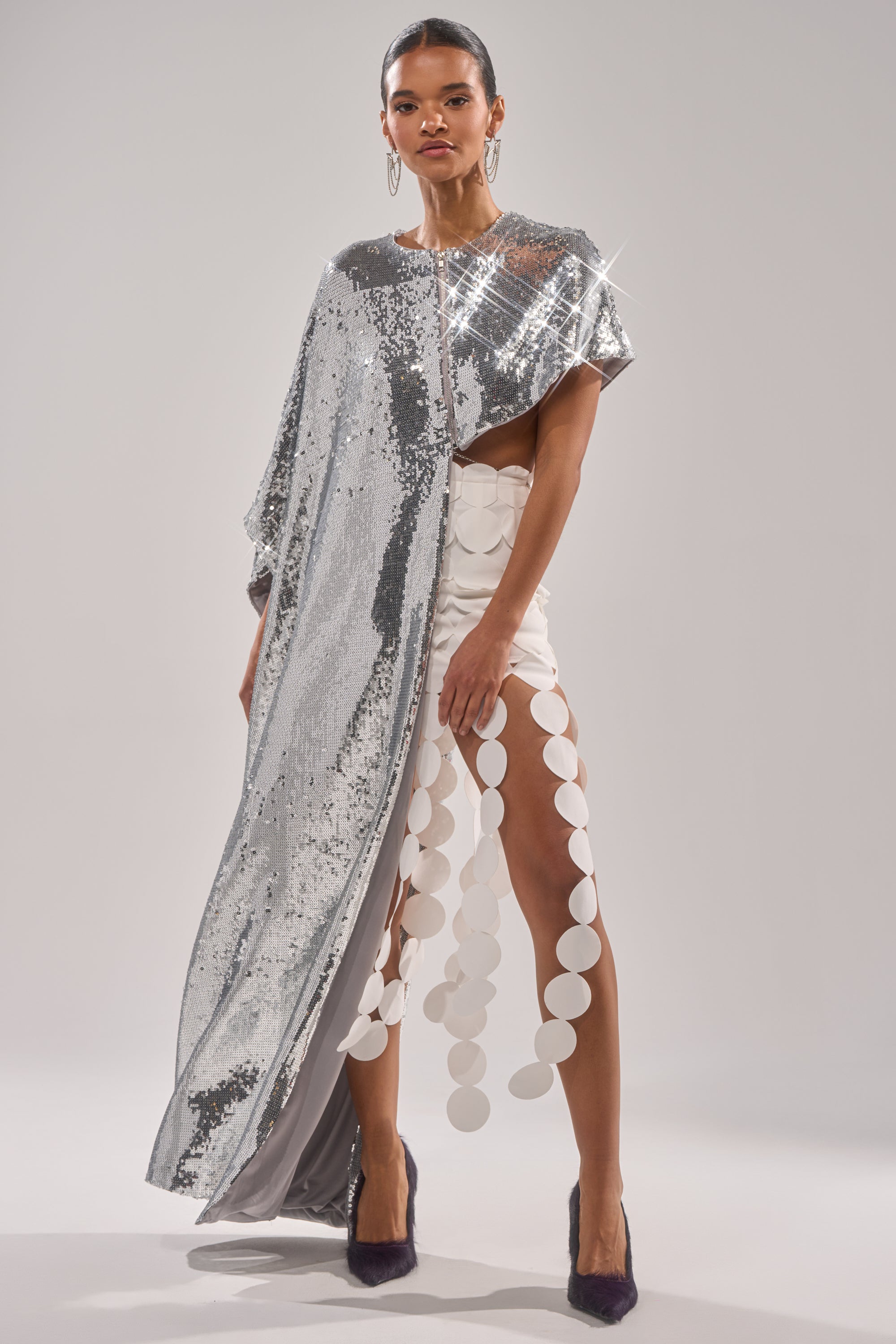 A woman wears the MIDDLE NAME IS DRAMA HIGH LOW BLOUSE, styled with hoop earrings and dark heels, posing against a plain background. The blouse features silver metallic sequins, an asymmetrical one-shoulder cut, and large white circular embellishments. 1