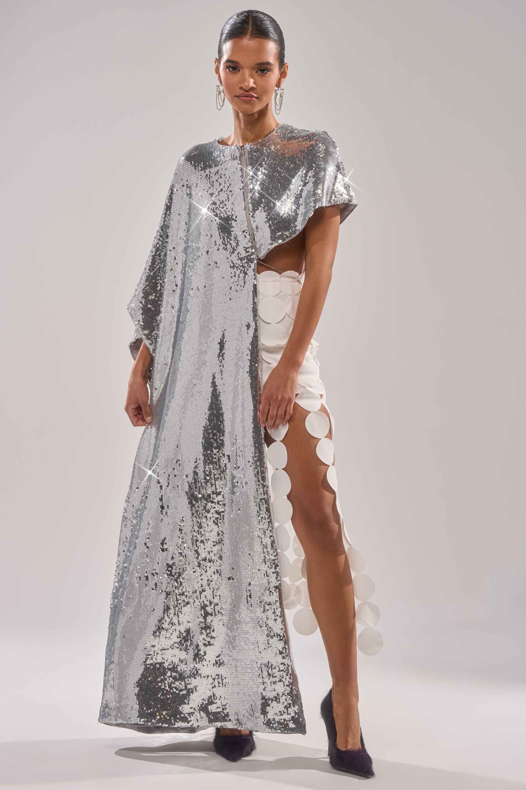 A woman wears the MIDDLE NAME IS DRAMA HIGH LOW BLOUSE—a shiny silver metallic sequin cape—over a white dress with large circular cutouts on one side and dark shoes, standing against a plain light gray background. 2