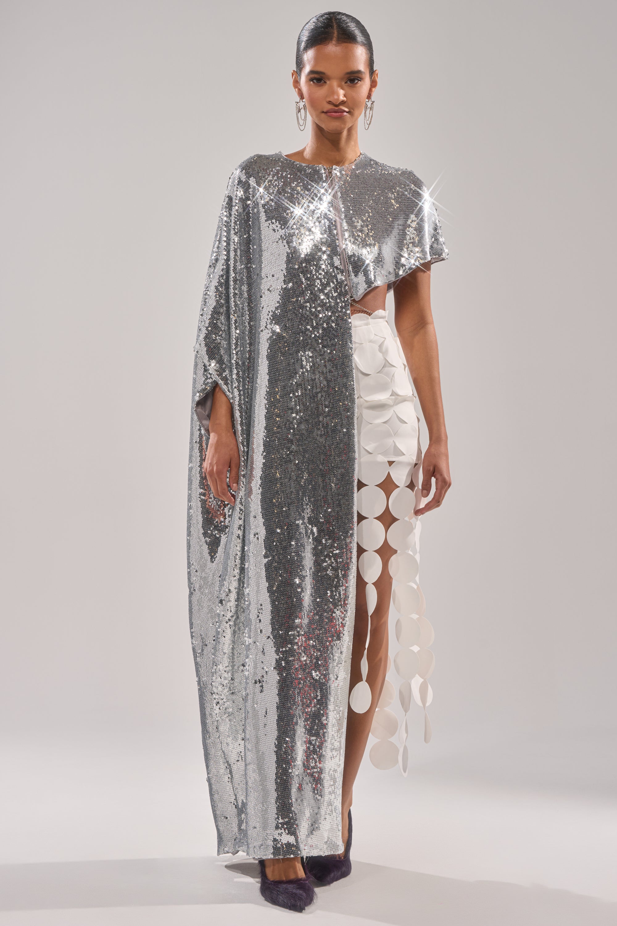 A woman poses in the shiny silver metallic "MIDDLE NAME IS DRAMA HIGH LOW BLOUSE" with a dramatic one-shoulder cape, paired with a white skirt featuring bold circular cutouts, set against a plain light background. 3