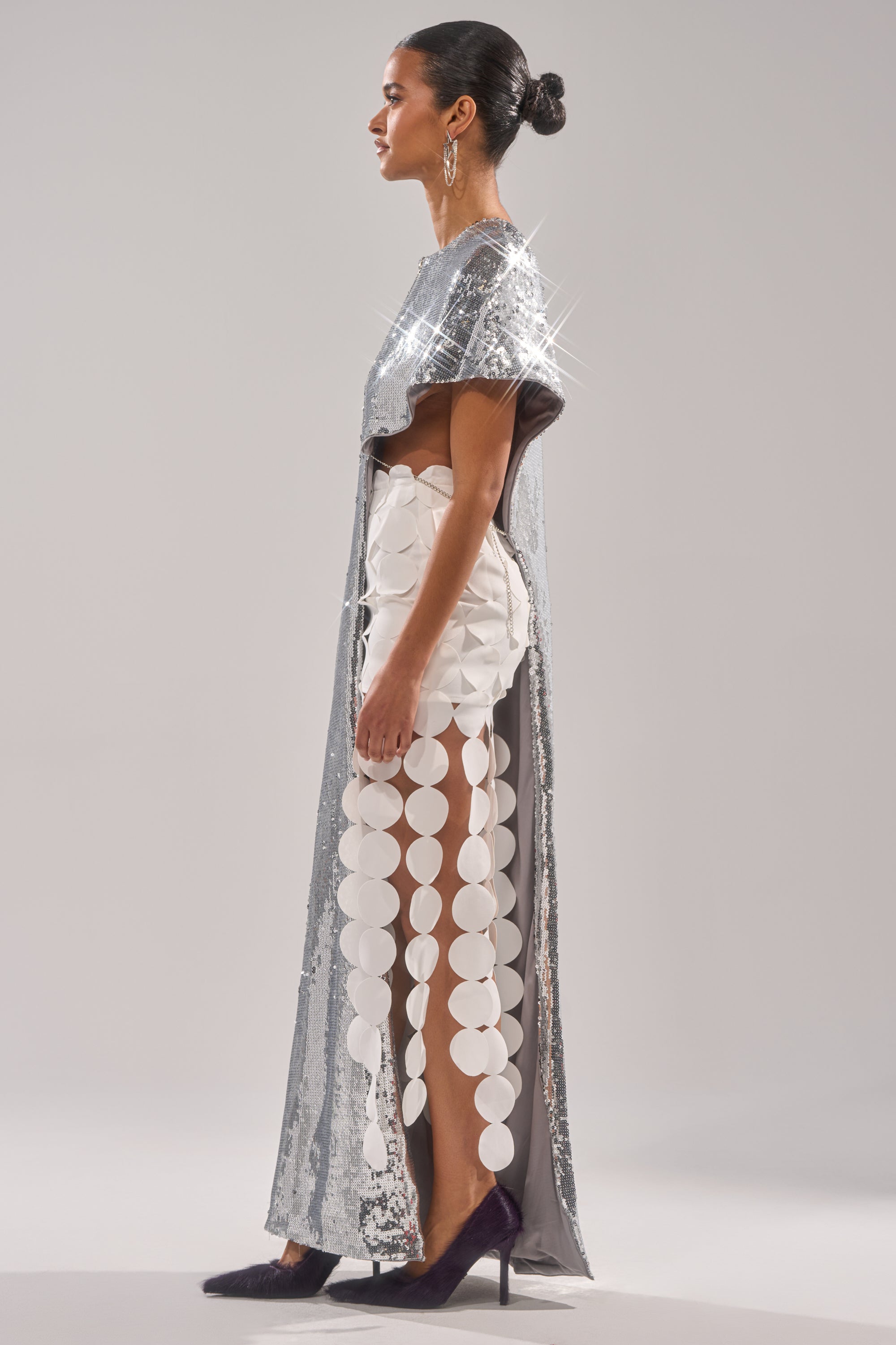 A woman stands in profile wearing the "Middle Name Is Drama High Low Blouse," styled with a cape-like top, large white circular cutouts on one side, a sleek bun, and pointed black heels. 4