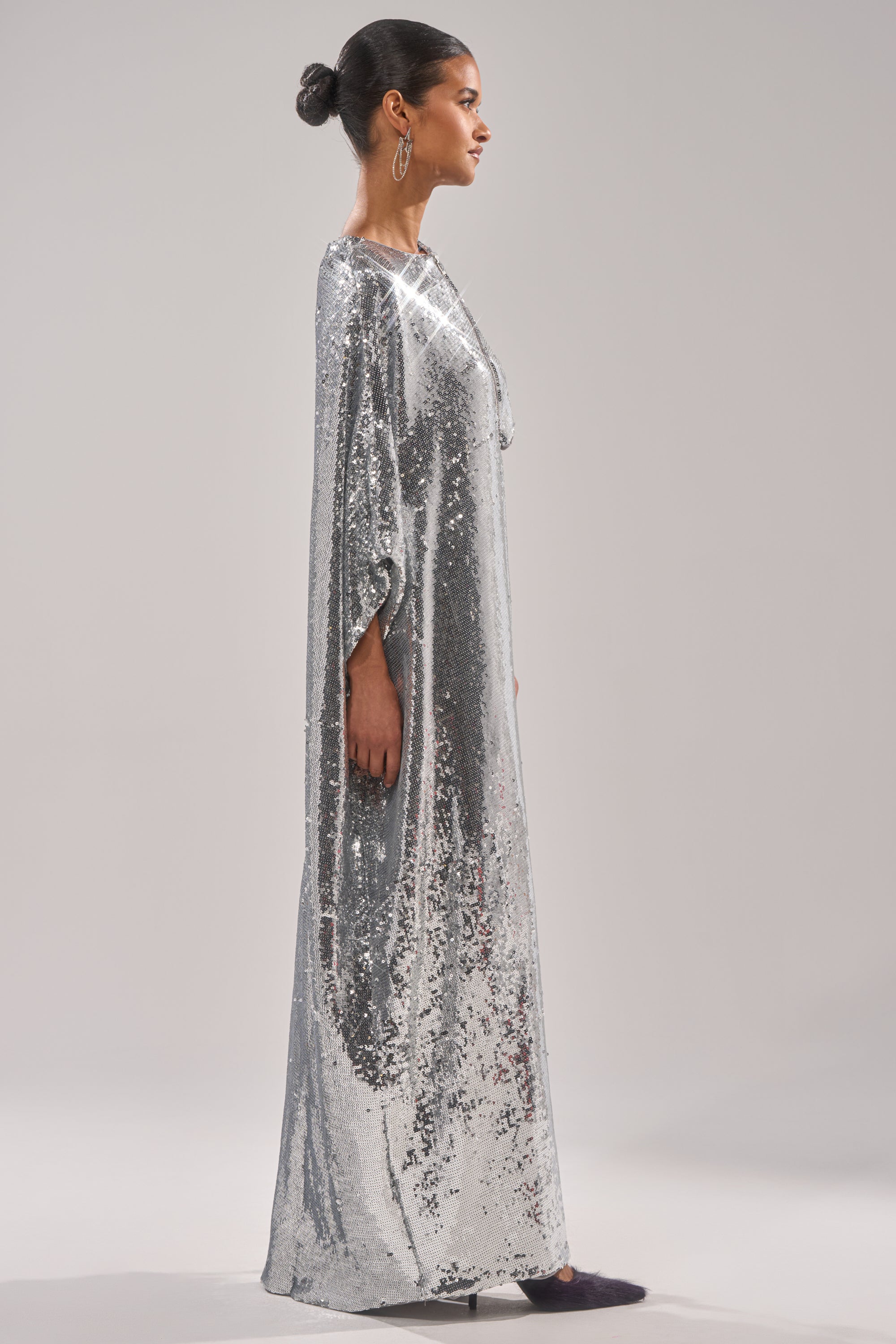 A woman stands in profile wearing the “MIDDLE NAME IS DRAMA HIGH LOW BLOUSE” with loose sleeves, her hair in a neat bun and accessorized with large hoop earrings, set against a plain, light background. 5