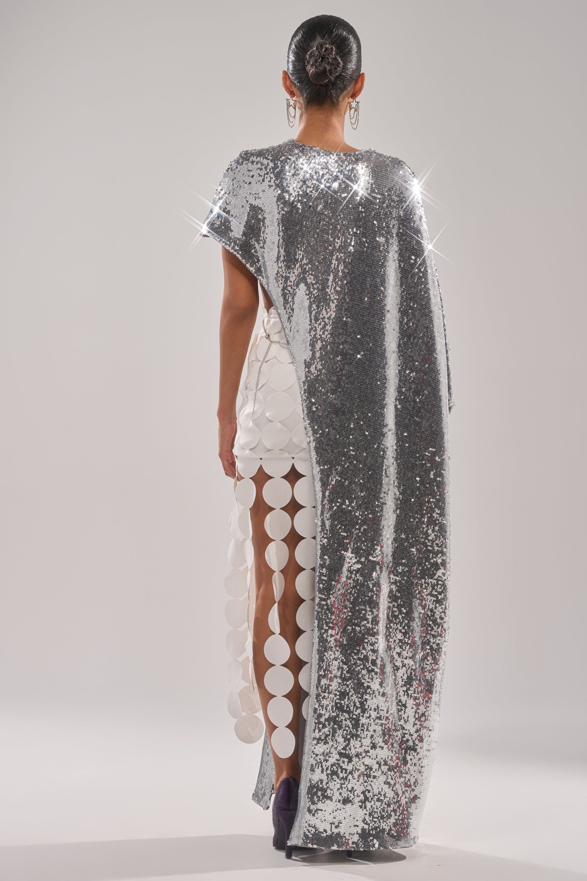 A model faces away wearing the MIDDLE NAME IS DRAMA HIGH LOW BLOUSE in shimmering silver sequins, draped over a white dress with bold circular cutouts. The look is finished with sparkling details and a sleek bun hairstyle. 6
