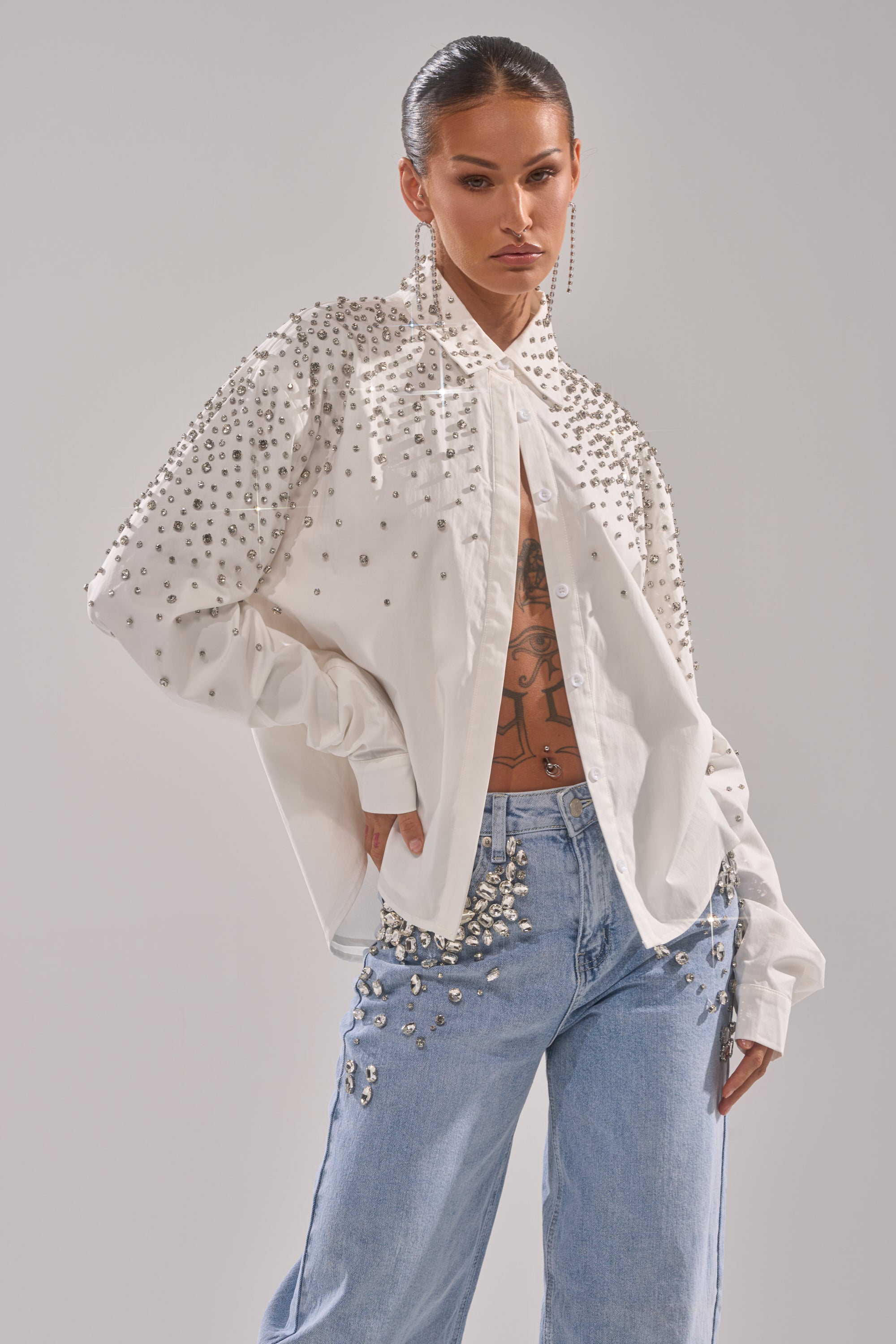 A model in the DIAMONDS FOREVER BUTTON DOWN and light blue jeans, both featuring silver studs. The look is styled with slicked-back hair, long earrings, and visible torso tattoos.
