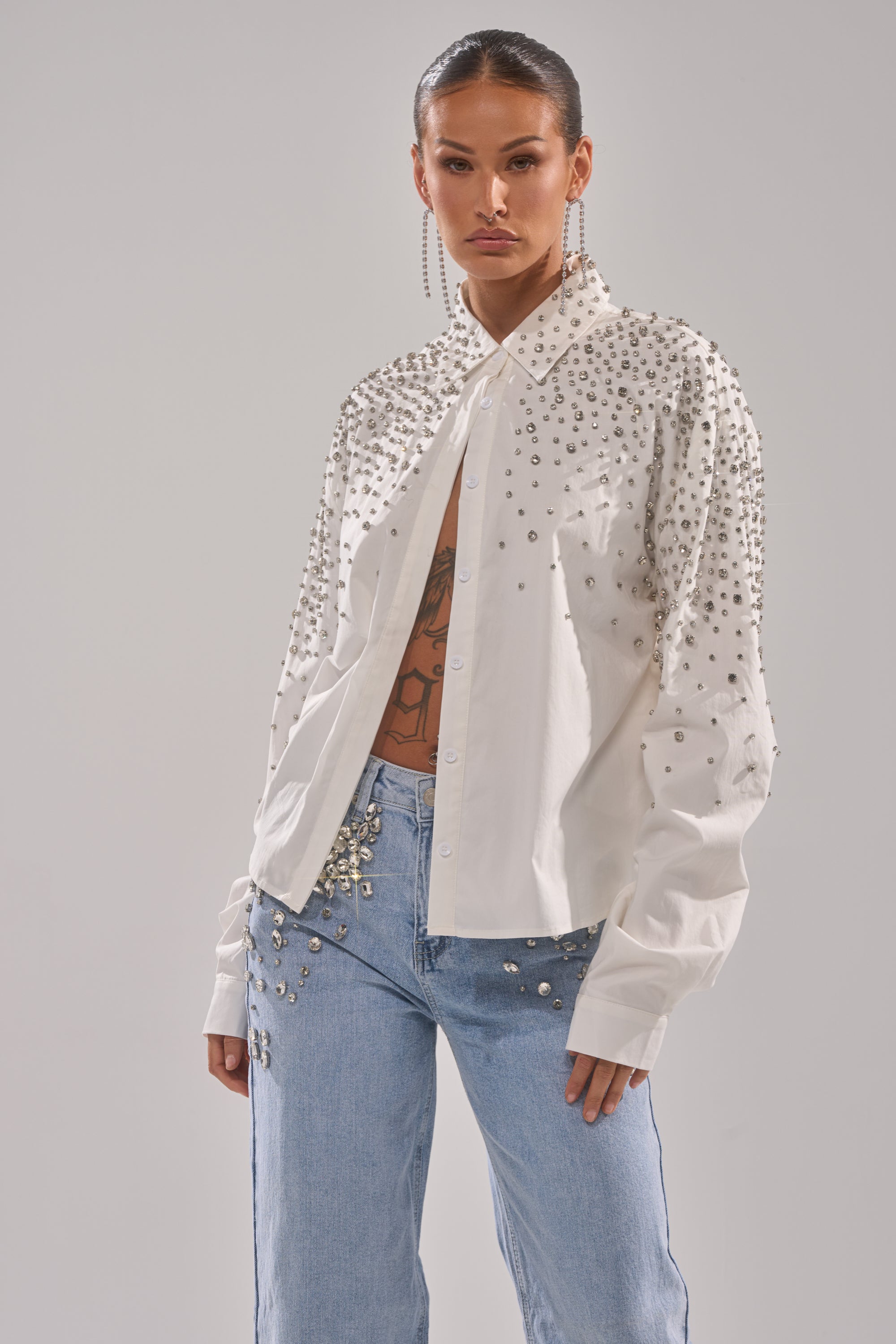 Wearing the DIAMONDS FOREVER BUTTON DOWN with light blue jeans, both featuring metallic studs, this bold look is finished with long earrings, slicked-back hair, and a glimpse of a chest tattoo beneath the slightly open shirt.