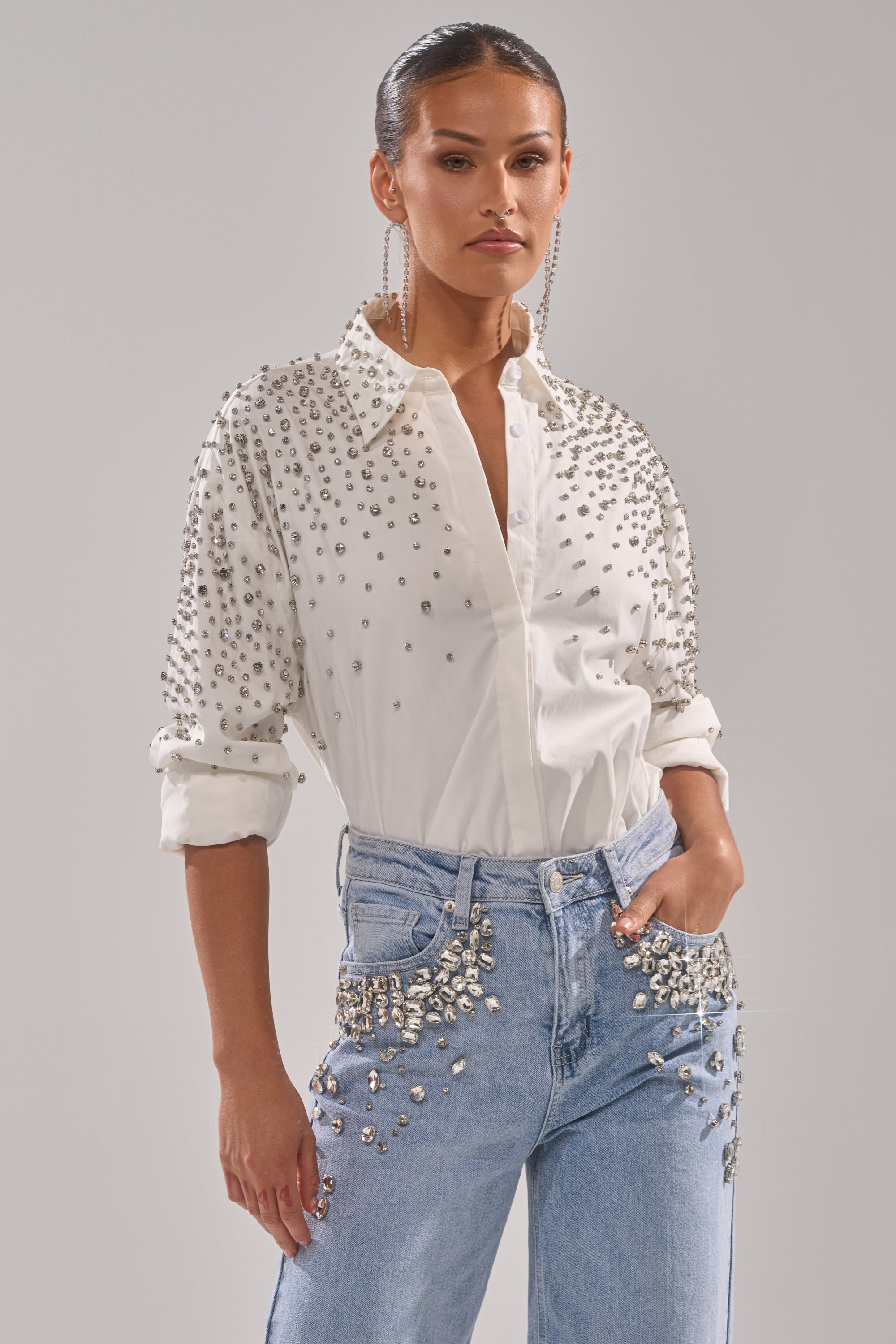 A woman exudes confidence in the DIAMONDS FOREVER BUTTON DOWN—a white, tailored shirt adorned with silver rhinestones—paired with studded light blue jeans, slicked-back hair, and long dangling earrings.