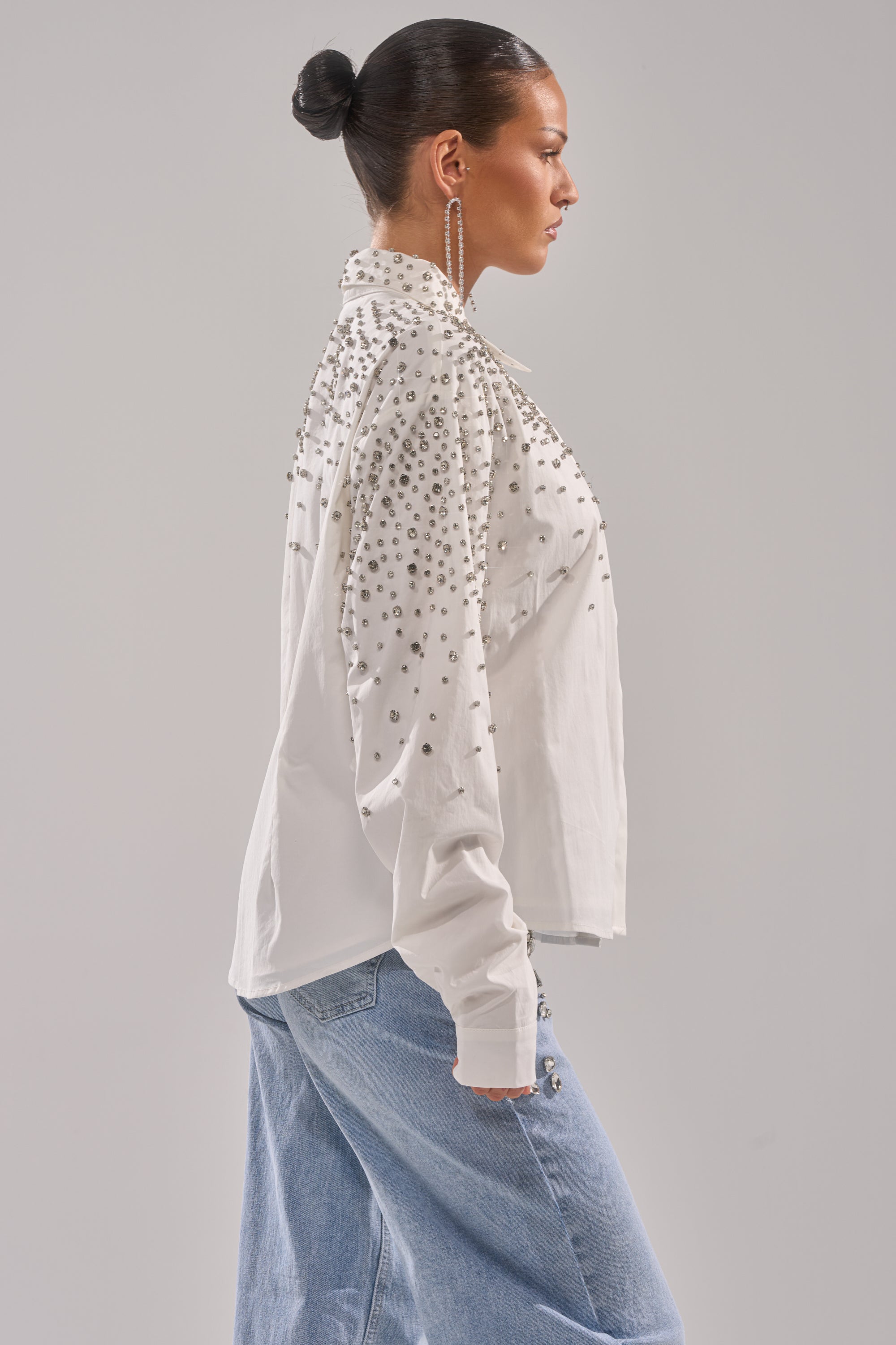 A woman with dark hair in a bun wears the DIAMONDS FOREVER BUTTON DOWN shirt with silver studs, long dangling earrings, and light blue jeans, standing in profile against a plain background.