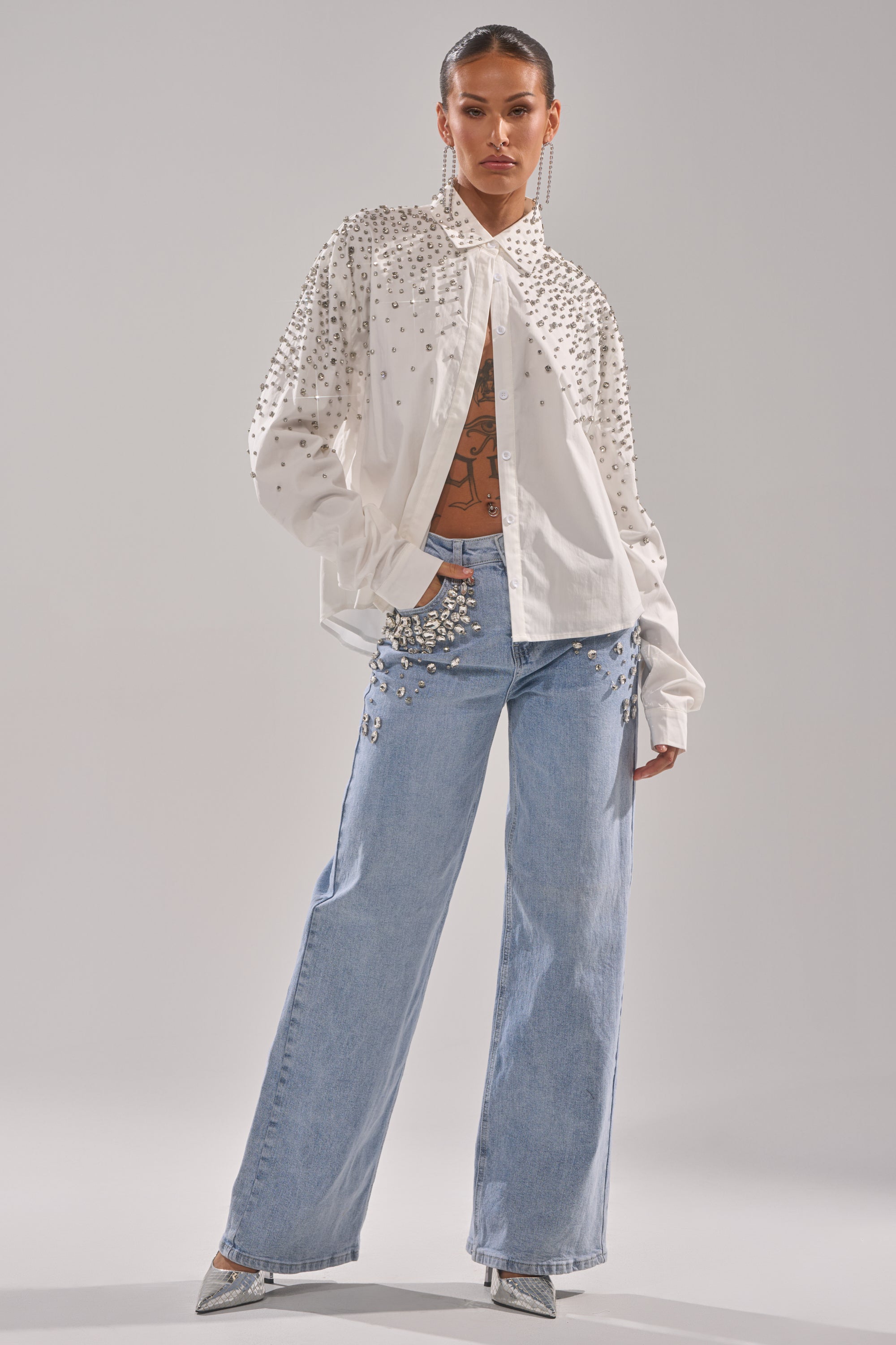 A person stands confidently in the DIAMONDS FOREVER BUTTON DOWN, paired with light blue wide-leg jeans with pearl details, silver pointed heels, and visible torso tattoos.