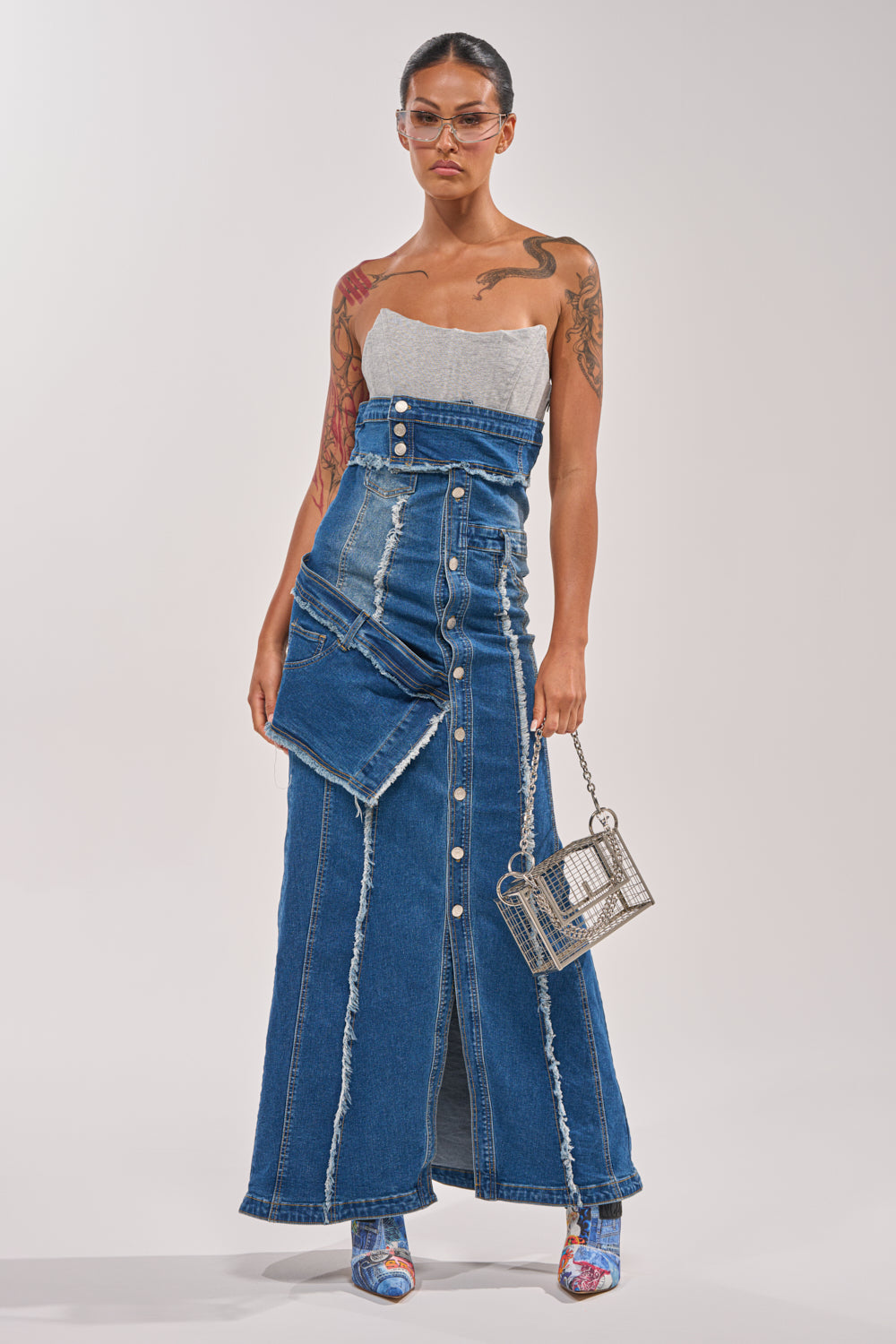 A woman wears the UP TO NO GOOD DISTRESSED DENIM MAXI DRESS with button details and visible seams, styling it with clear glasses, blue heeled sandals, and a metallic square handbag against a plain backdrop for a standout festival look. 1