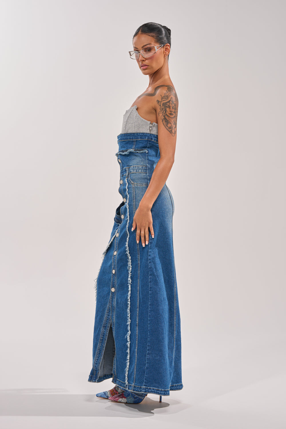 A person models the UP TO NO GOOD DISTRESSED DENIM MAXI DRESS, featuring a strapless corset top, button-down front, frayed edges, and high slit. They accessorize with clear glasses and high heels against a plain light background. 3