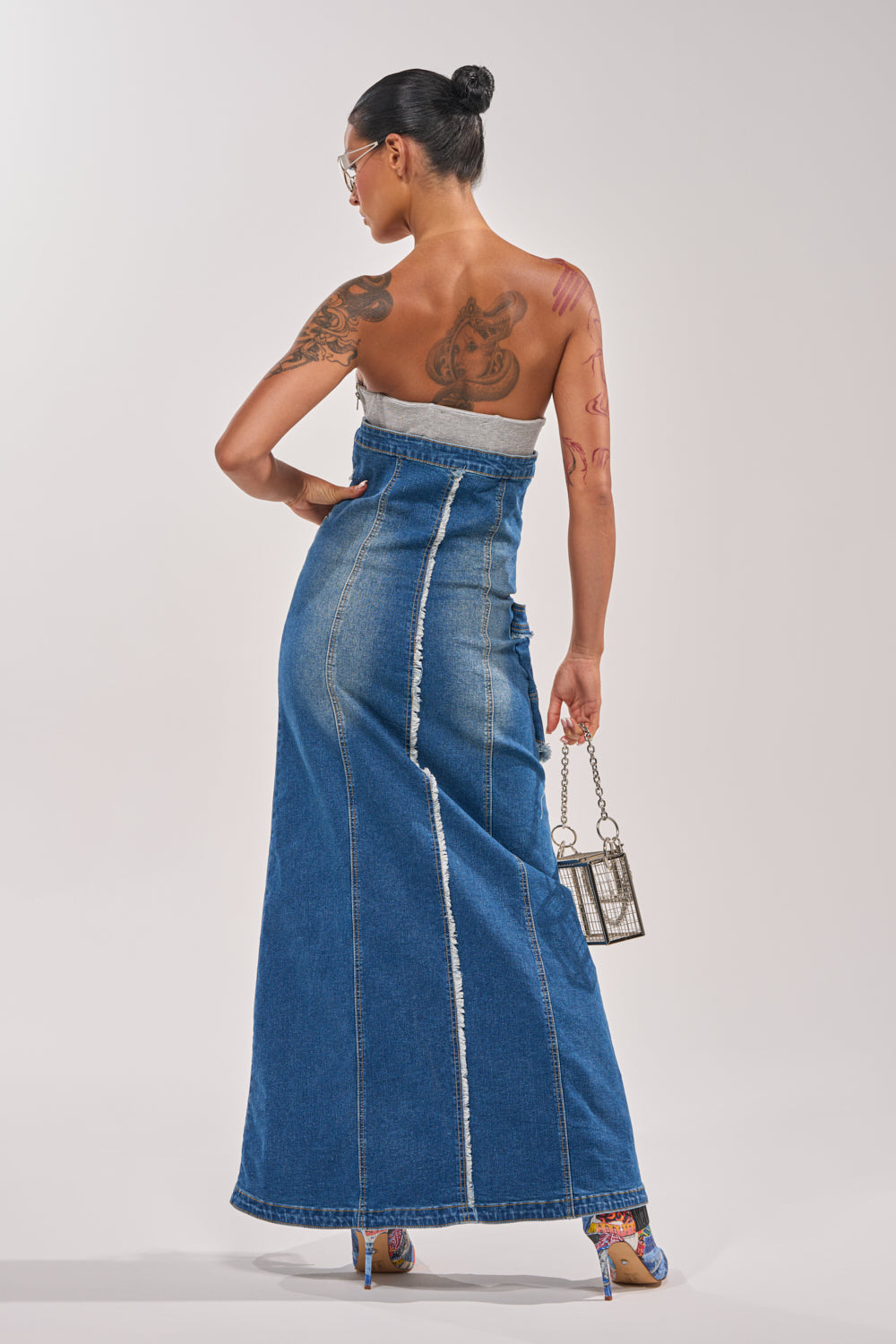 A woman with a bun hairstyle and tattoos poses with her back to the camera in the UP TO NO GOOD DISTRESSED DENIM MAXI DRESS, styled with colorful heels and a metallic handbag—an ideal festival outfit against a plain backdrop. 4
