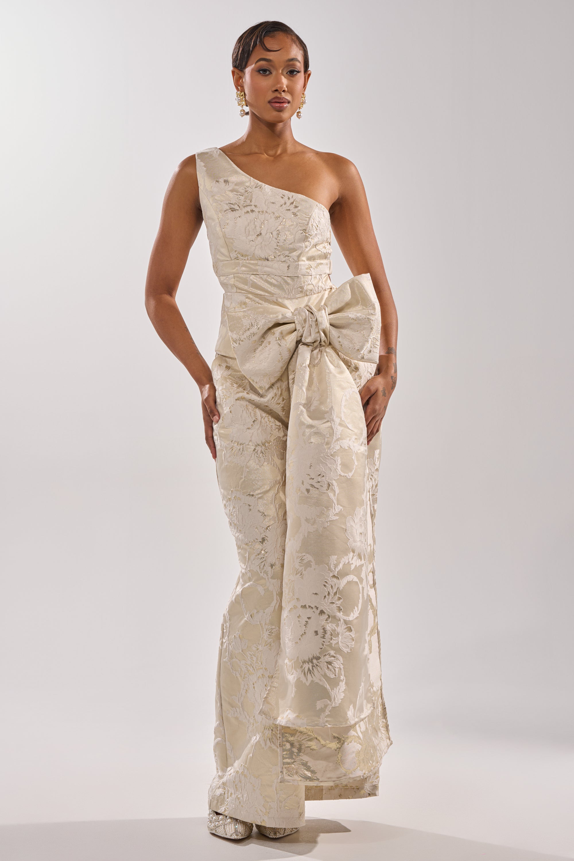 A woman models the DRESSED TO IMPRESS BLOUSE, featuring elegant textured jacquard, a one-shoulder design, a large waist bow, and a peplum hem. She pairs it with statement earrings against a simple light background.