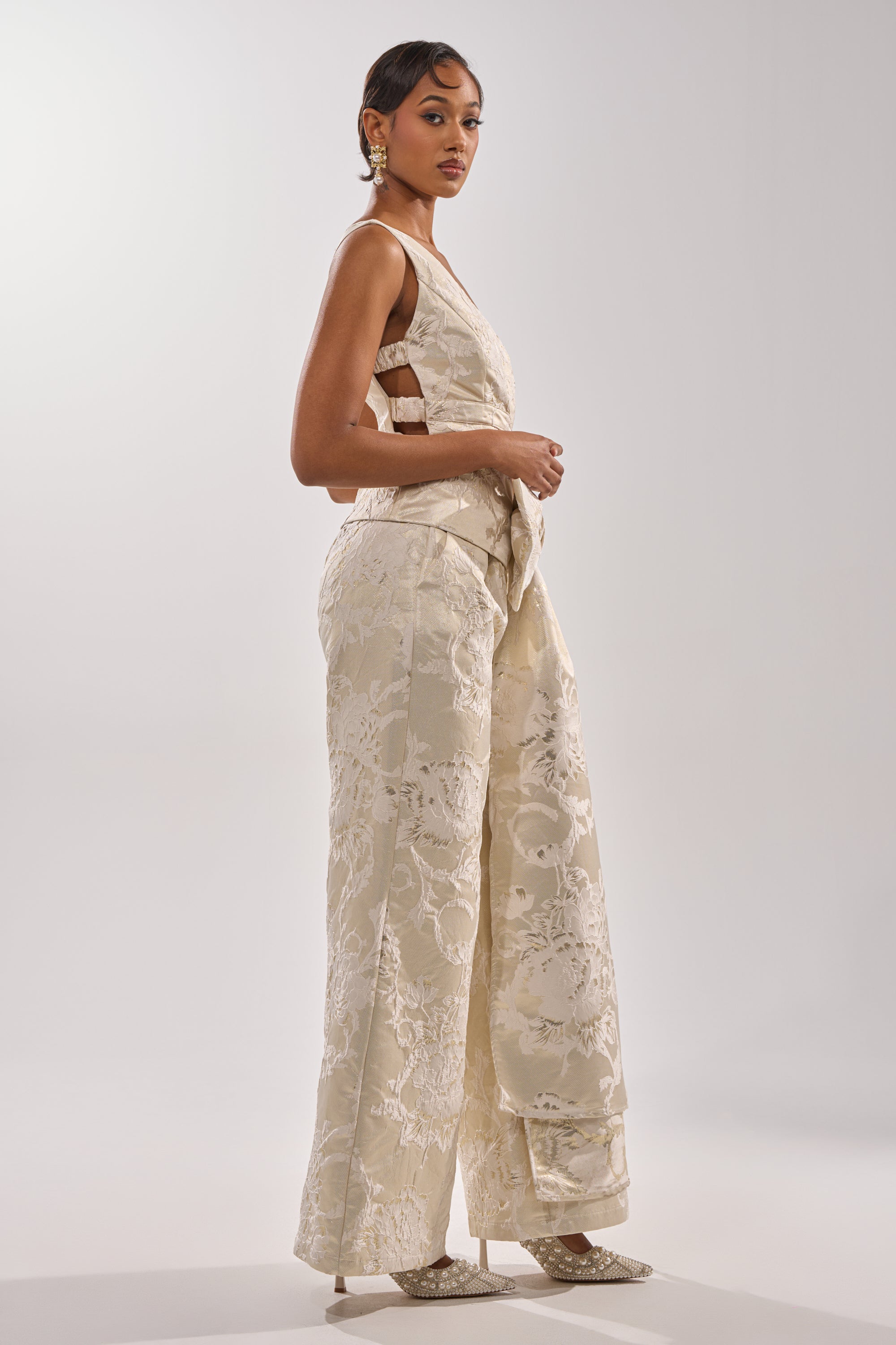 A woman in the DRESSED TO IMPRESS BLOUSE, a sleeveless cream jacquard top with side cutouts, pairs it with matching wide-leg pants, statement earrings, pointed heels, and wears her hair short against a plain background.