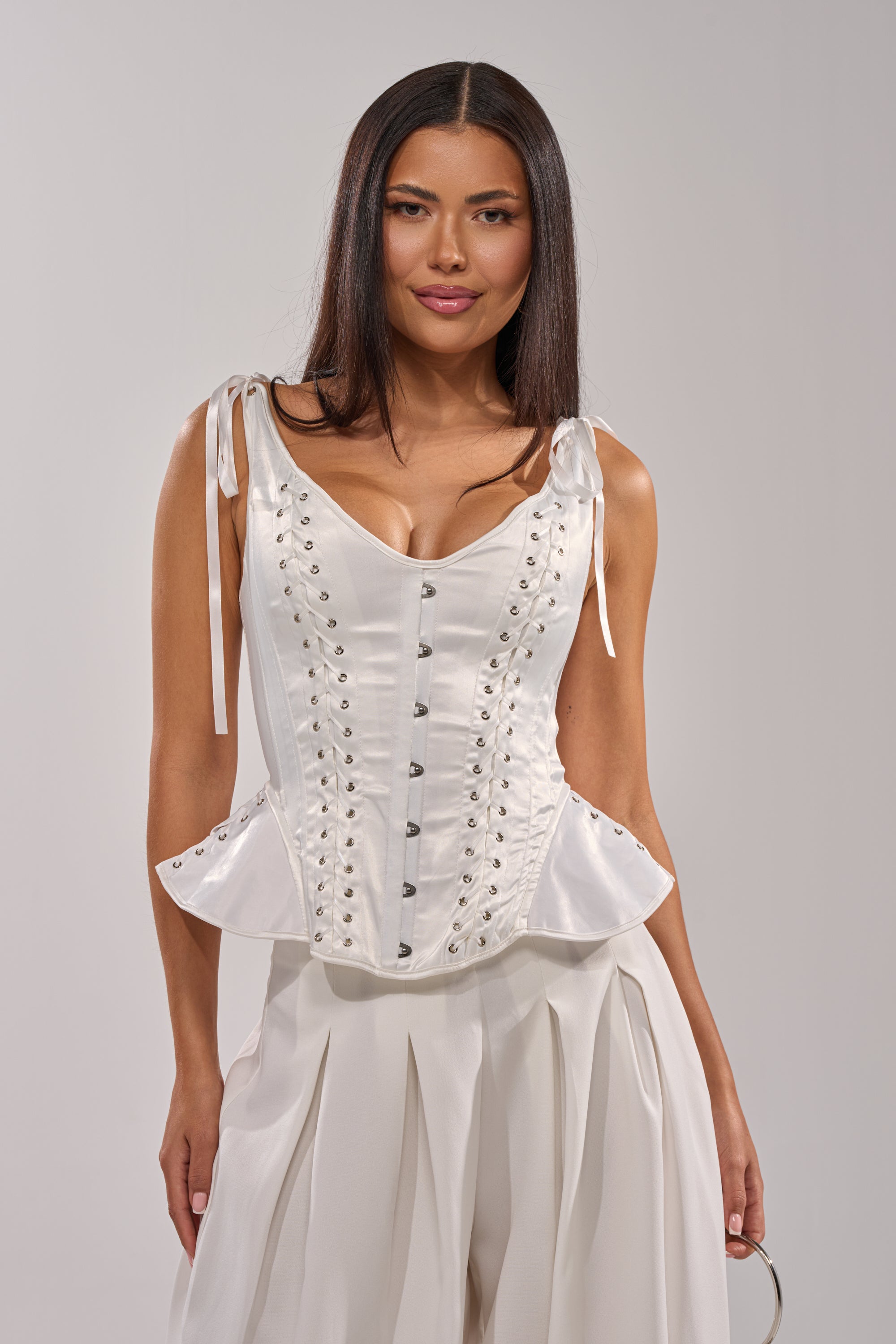 A woman with long dark hair models the FEELING THE VIBES CORSET, a white lace-up top with decorative buttons and sculptural hips, paired with matching wide-leg pleated pants against a plain light background. 4