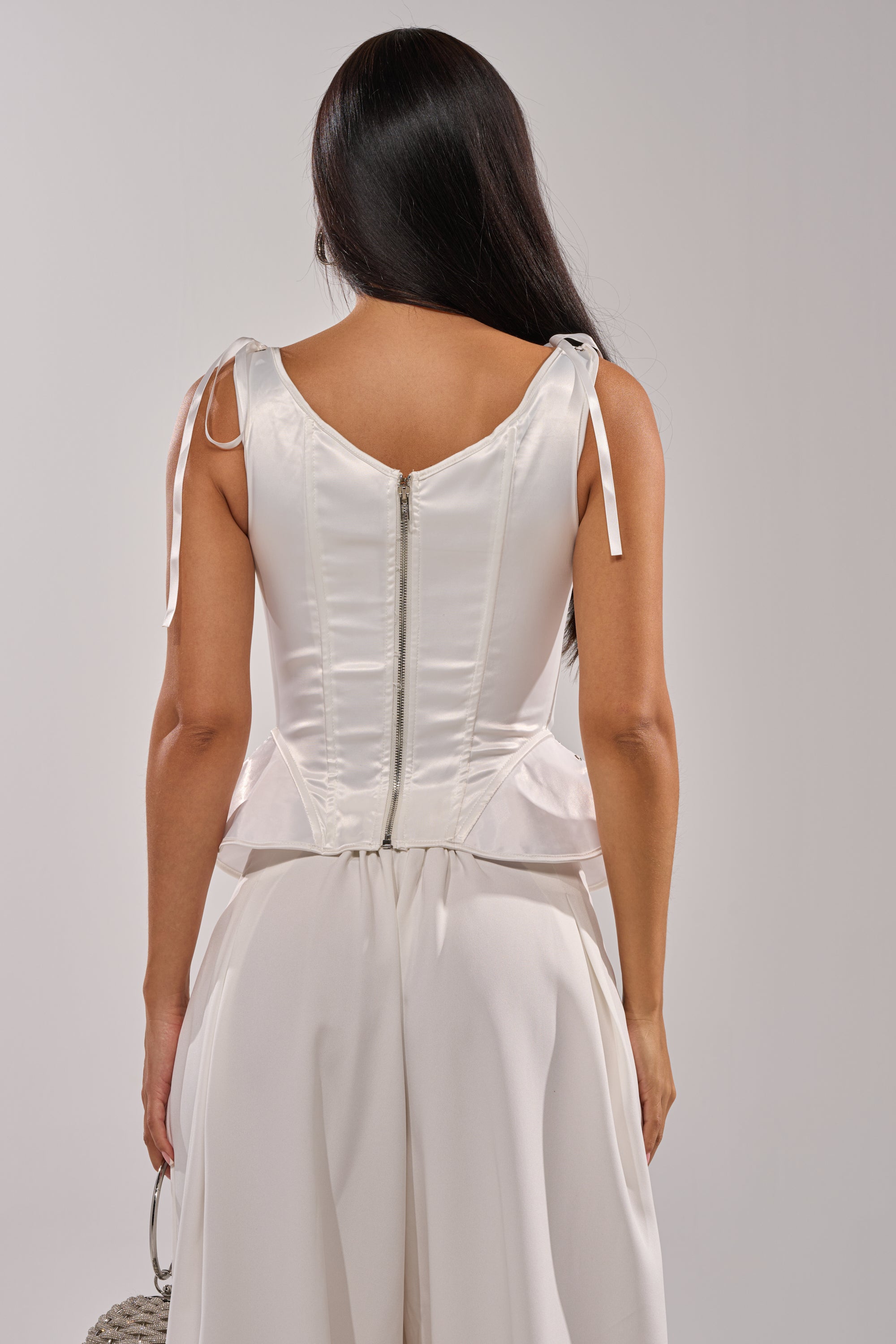 A woman with long dark hair wears the FEELING THE VIBES CORSET—a sleeveless white lace-up top with shoulder ties and a visible back zipper—paired with loose white pants, standing with her back to the camera against a light gray background. 6