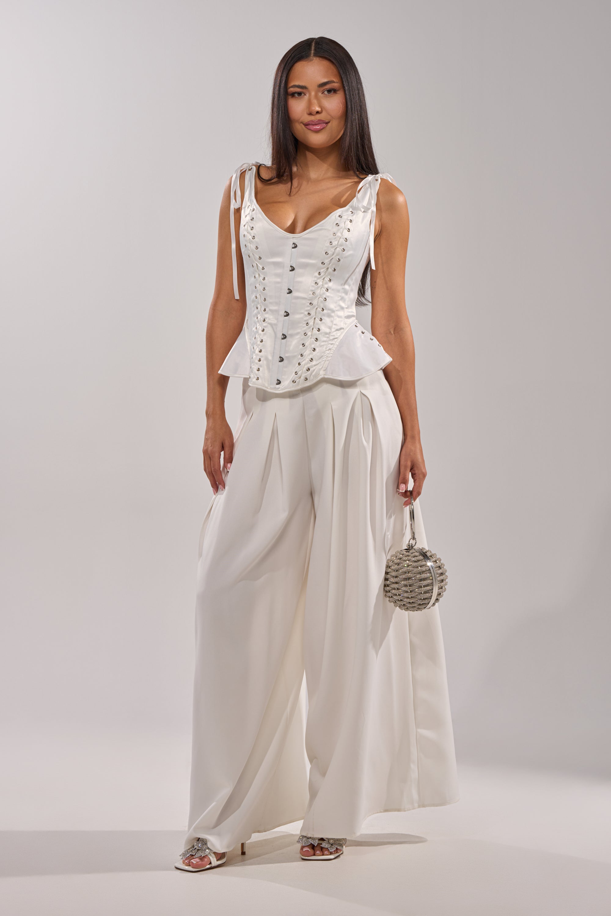 A woman stands against a plain background wearing the FEELING THE VIBES CORSET and wide-leg trousers, holding a small round silver handbag and wearing open-toe heels. 7