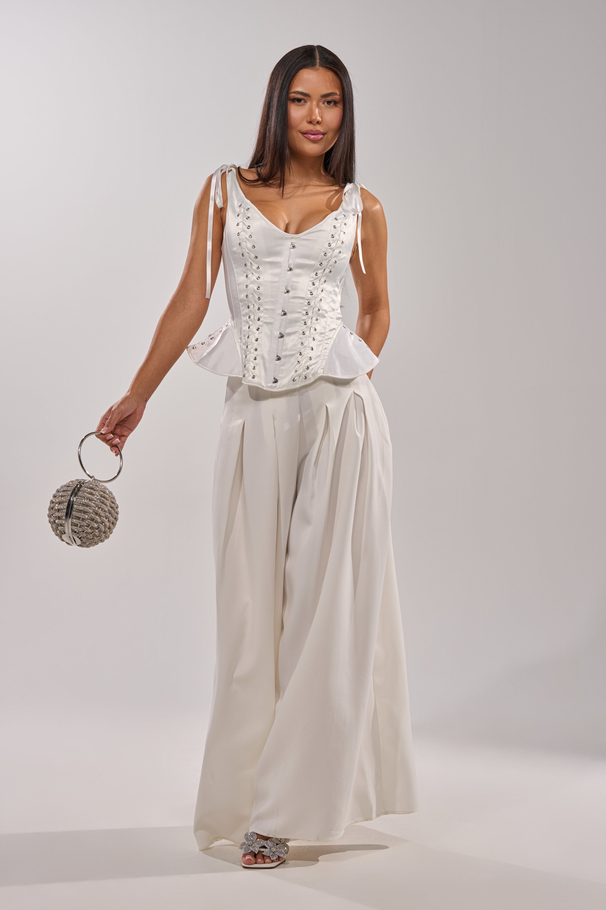 A woman poses in the FEELING THE VIBES CORSET and wide-leg white trousers, holding a round silver handbag. She has long straight hair, open-toed shoes, and sculptural hips against a plain light background. 8