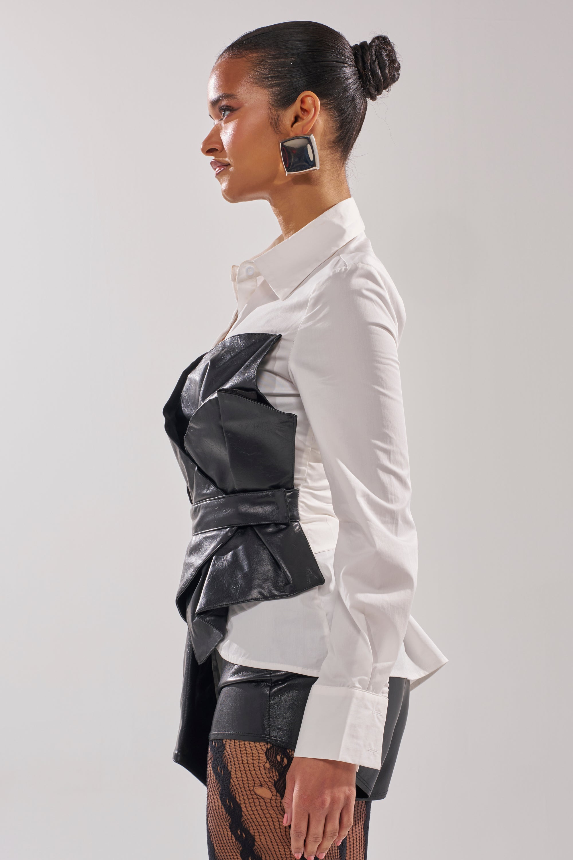 A woman in profile against a plain background wears the IS THIS CRAZY ENOUGH BLOUSE under a black faux leather overlay, paired with black shorts, patterned tights, and large square earrings. Her hair is slicked back in a neat bun. 3