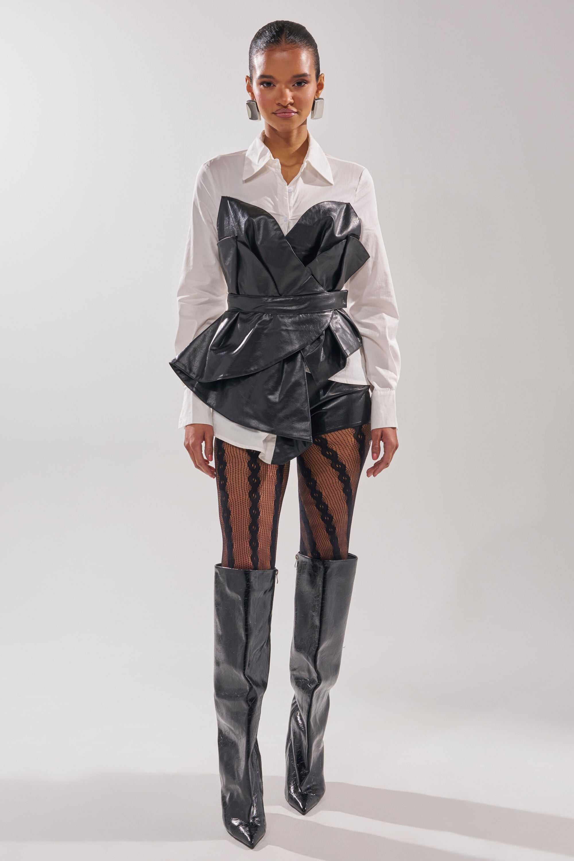 A woman faces forward in the IS THIS CRAZY ENOUGH BLOUSE, a sculptural faux leather top layered over a white collared shirt. She pairs it with patterned black tights and knee-high black boots against a plain, light background. 6