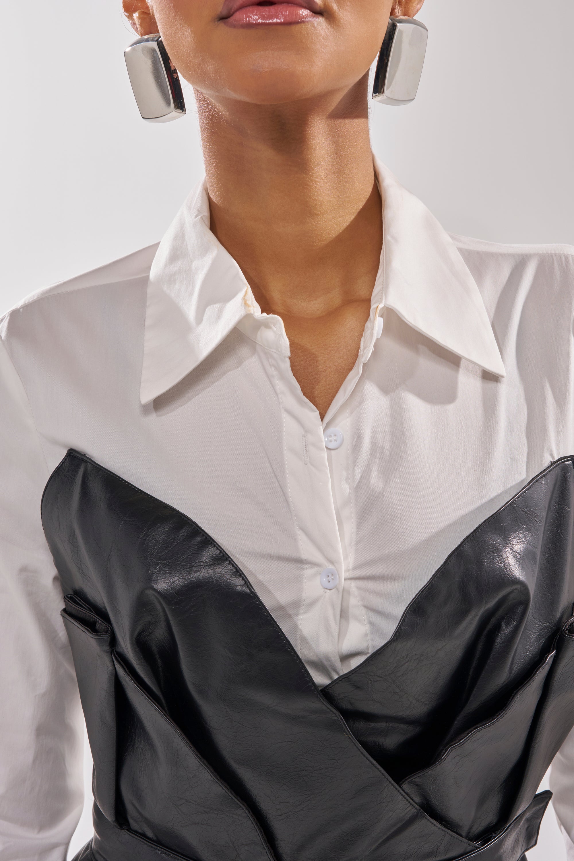A person wears the IS THIS CRAZY ENOUGH BLOUSE, a white sculptural top layered with a black faux leather bustier and large geometric silver earrings; only their lower face and upper torso are visible. 8