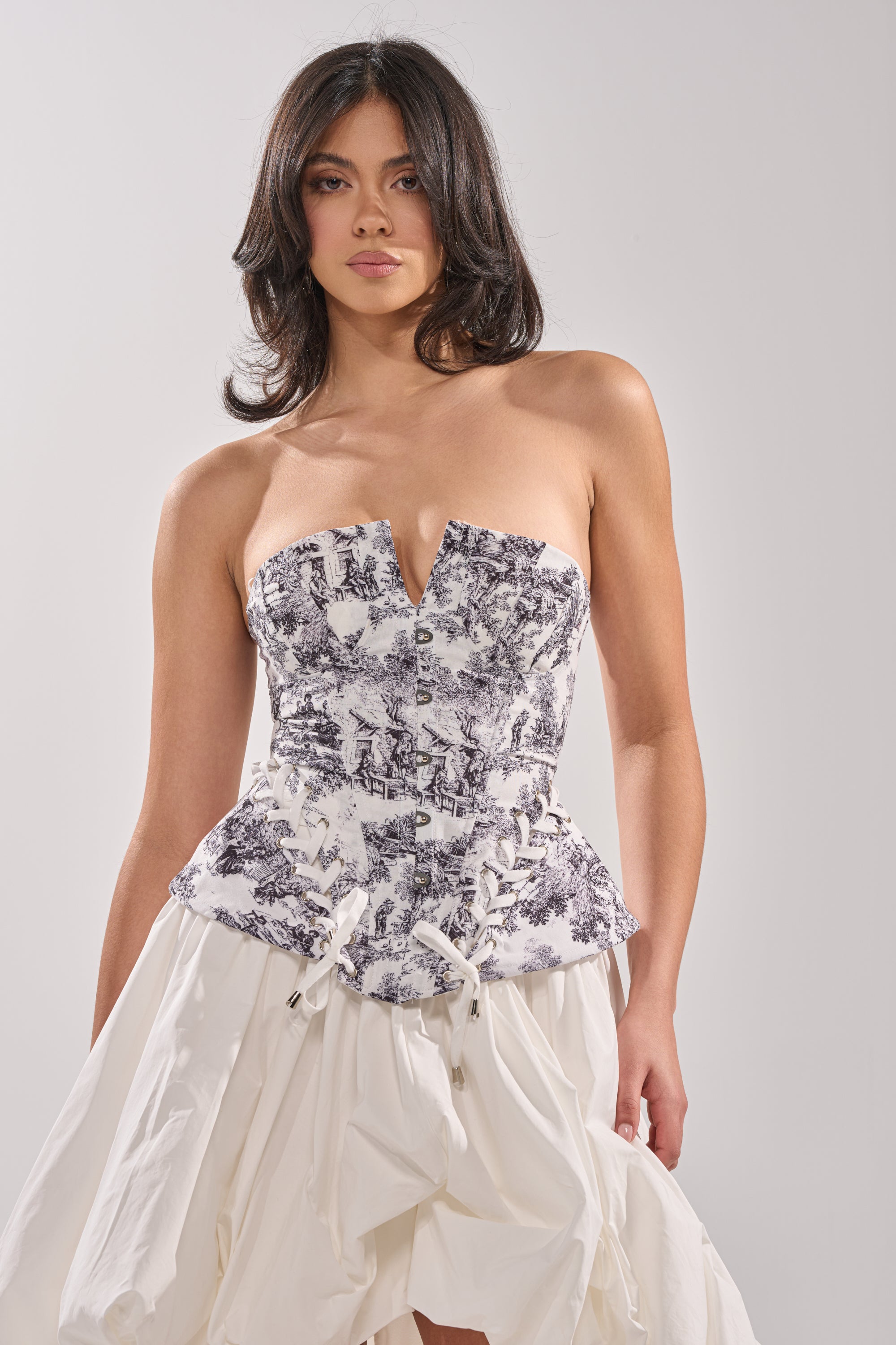 A woman with wavy dark hair wears the DREAMER GIRL CORSET, a strapless black and white toile peplum top, paired with a voluminous white skirt, posing against a plain light gray background. 0
