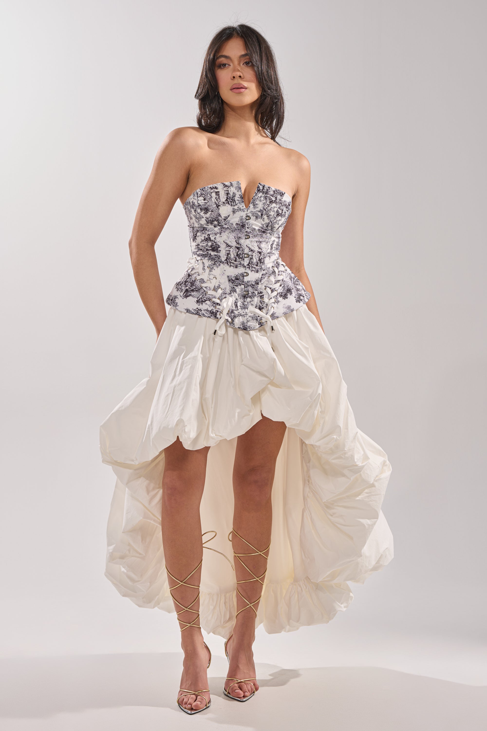 A woman wears the DREAMER GIRL CORSET, featuring a fitted toile print bodice, paired with a voluminous high-low white skirt. She completes her look with strappy heels and loose, wavy hair against a light background. 4