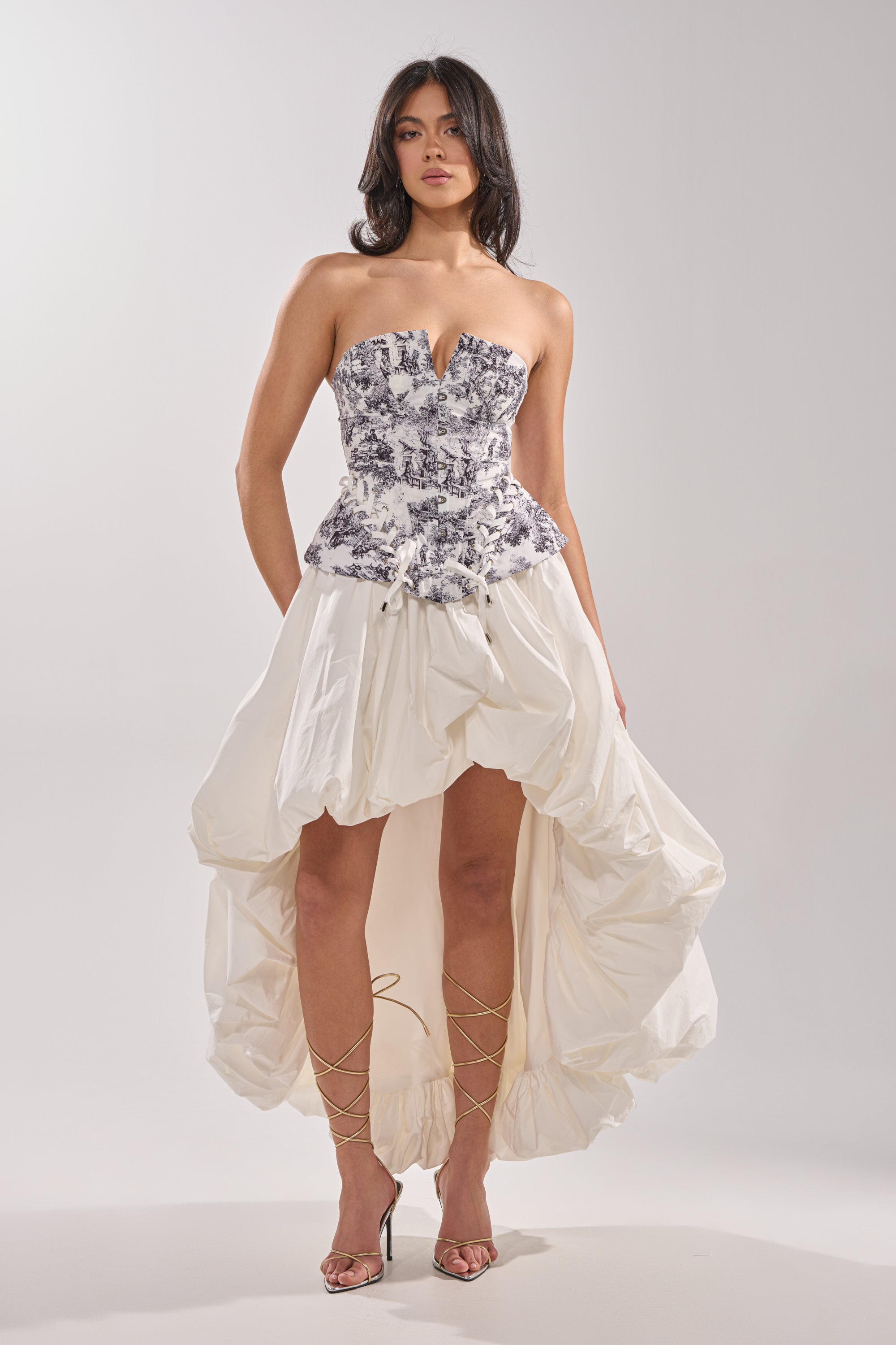 A woman wears the DREAMER GIRL CORSET—a strapless black-and-white toile print bodice—paired with a voluminous high-low white skirt and strappy gold heels. 5