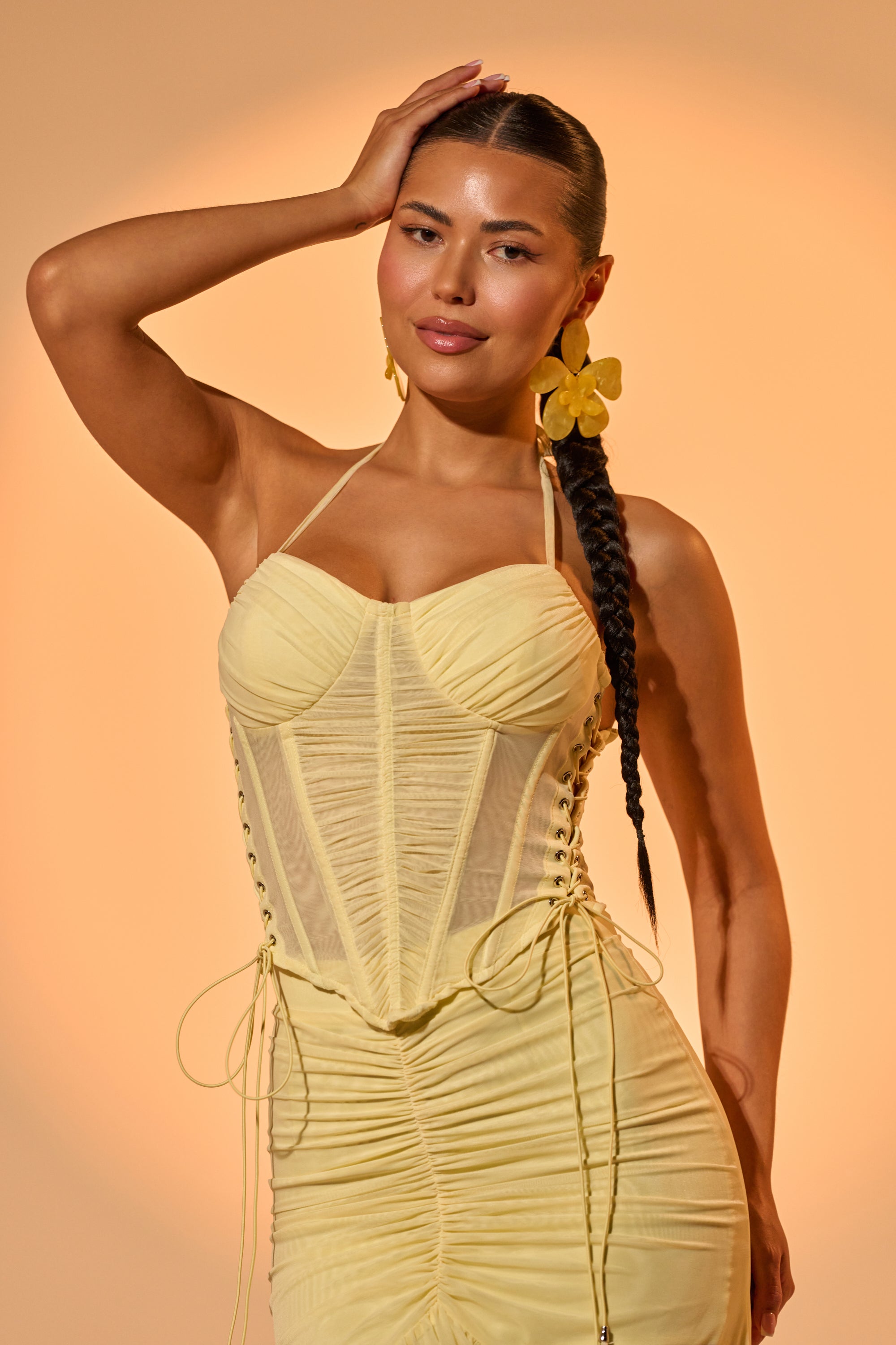 A woman with dark hair in a long braid poses in the VACA VIBES MESH CORSET, a pale yellow dress with ruched bust cups and a corset-style bodice. She accessorizes with large yellow flower earrings against a warm beige background. 1