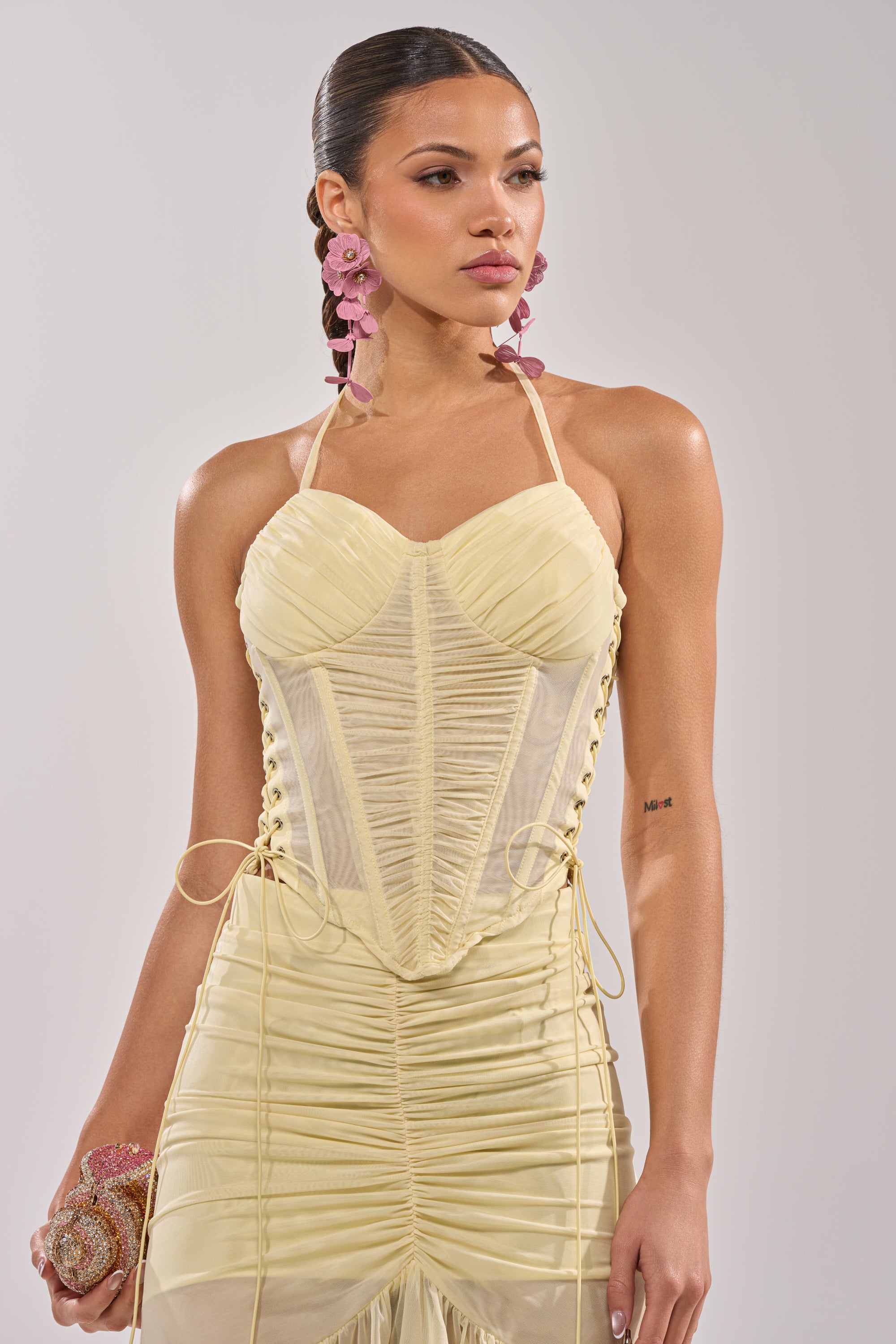 A woman wears a pale yellow dress featuring the VACA VIBES MESH CORSET bodice with ruched bust cups, paired with a beaded purse, large pink floral earrings, and slicked-back braided hair for an elegant look. 4