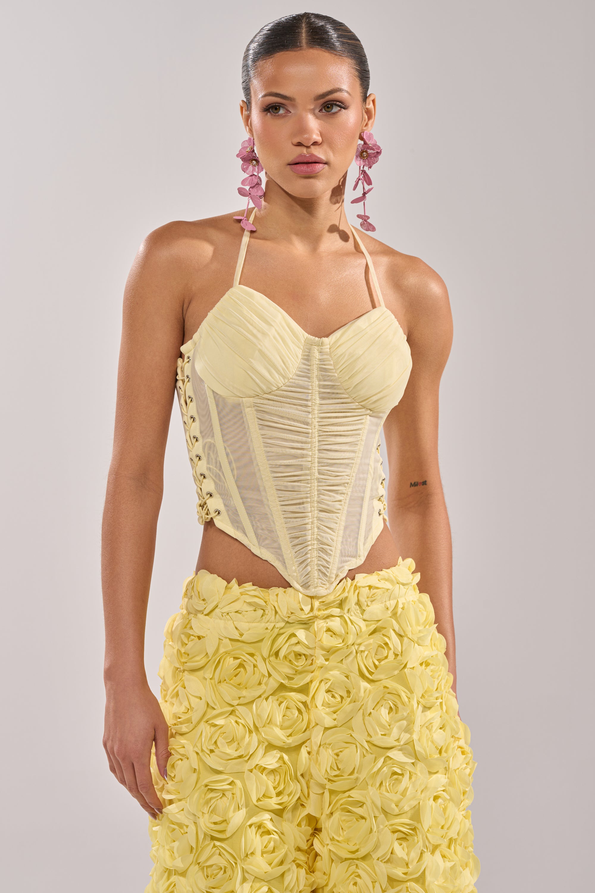 A woman wears a pale yellow, fitted VACA VIBES MESH CORSET with halter straps and ruched bust cups, paired with high-waisted pants featuring 3D rosette textures. She completes her look with pink statement earrings and slicked-back hair. 0