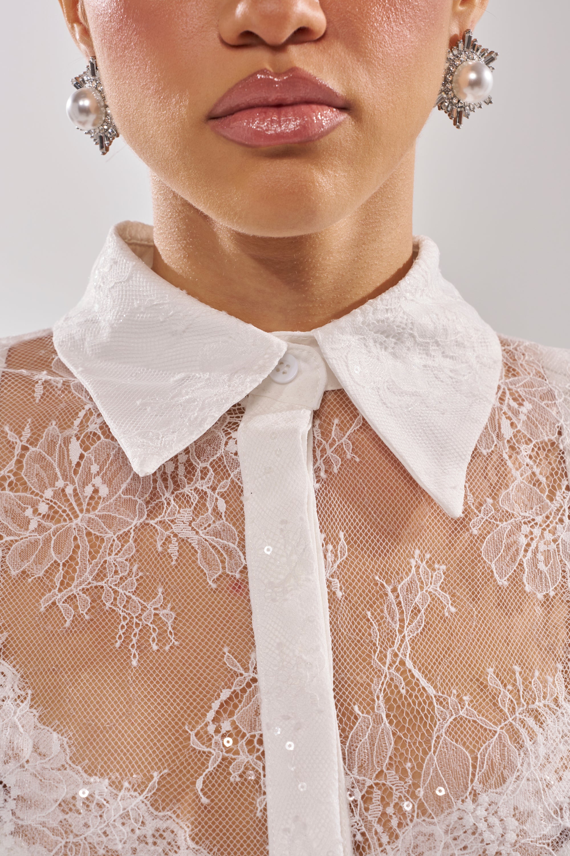 A woman wears the LADY LOVER LACE BUTTON DOWN, a sheer blouse with a solid white collar and tie, floral embroidery, and sequin details. Pearl earrings accent her look; only her lips, chin, and lower face are visible. 10