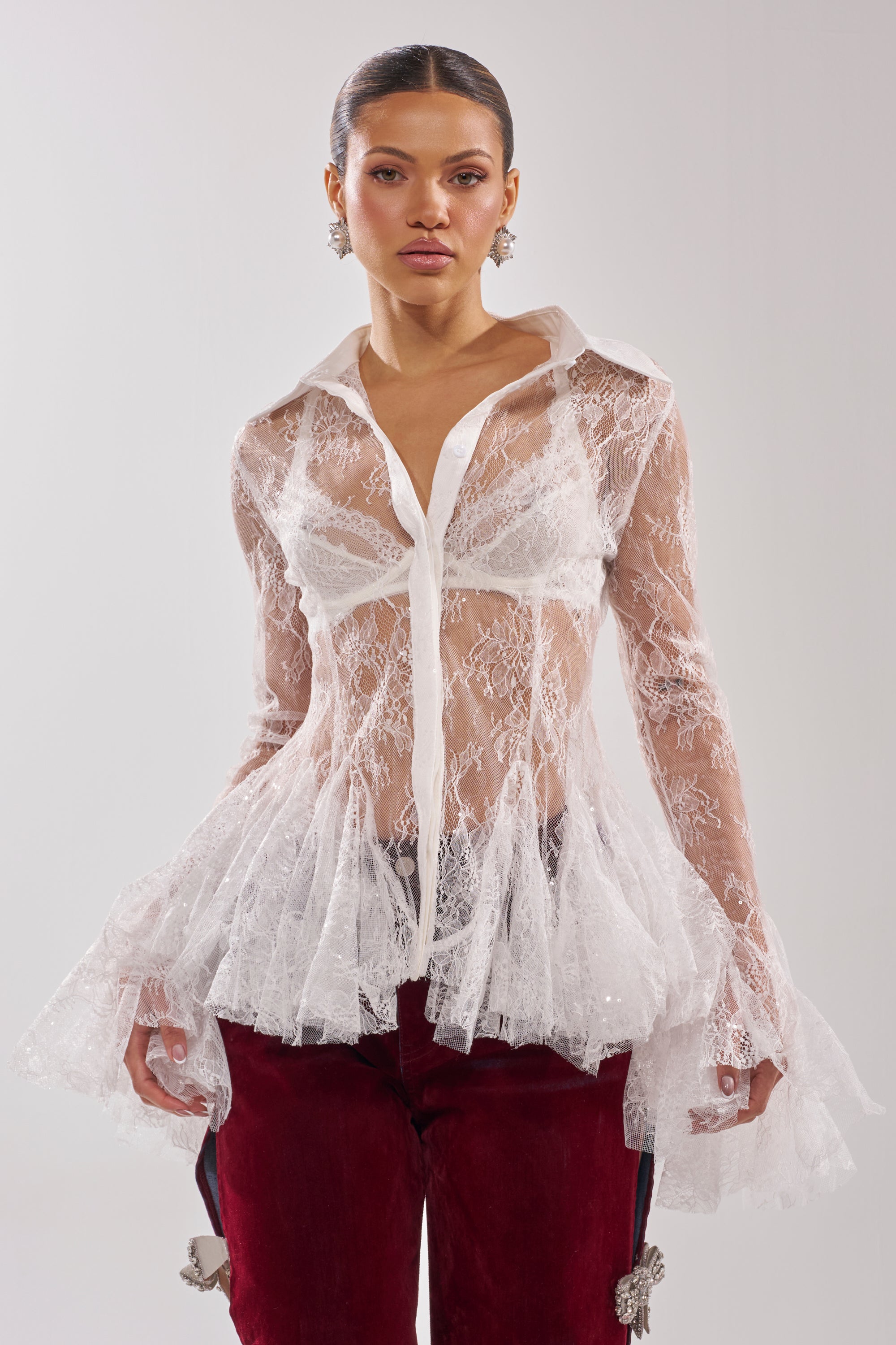 A woman models the LADY LOVER LACE BUTTON DOWN blouse, featuring sheer lace with a flared hem and collar, paired with high-waisted red velvet pants. She stands against a plain light background, confidently facing the camera. 2