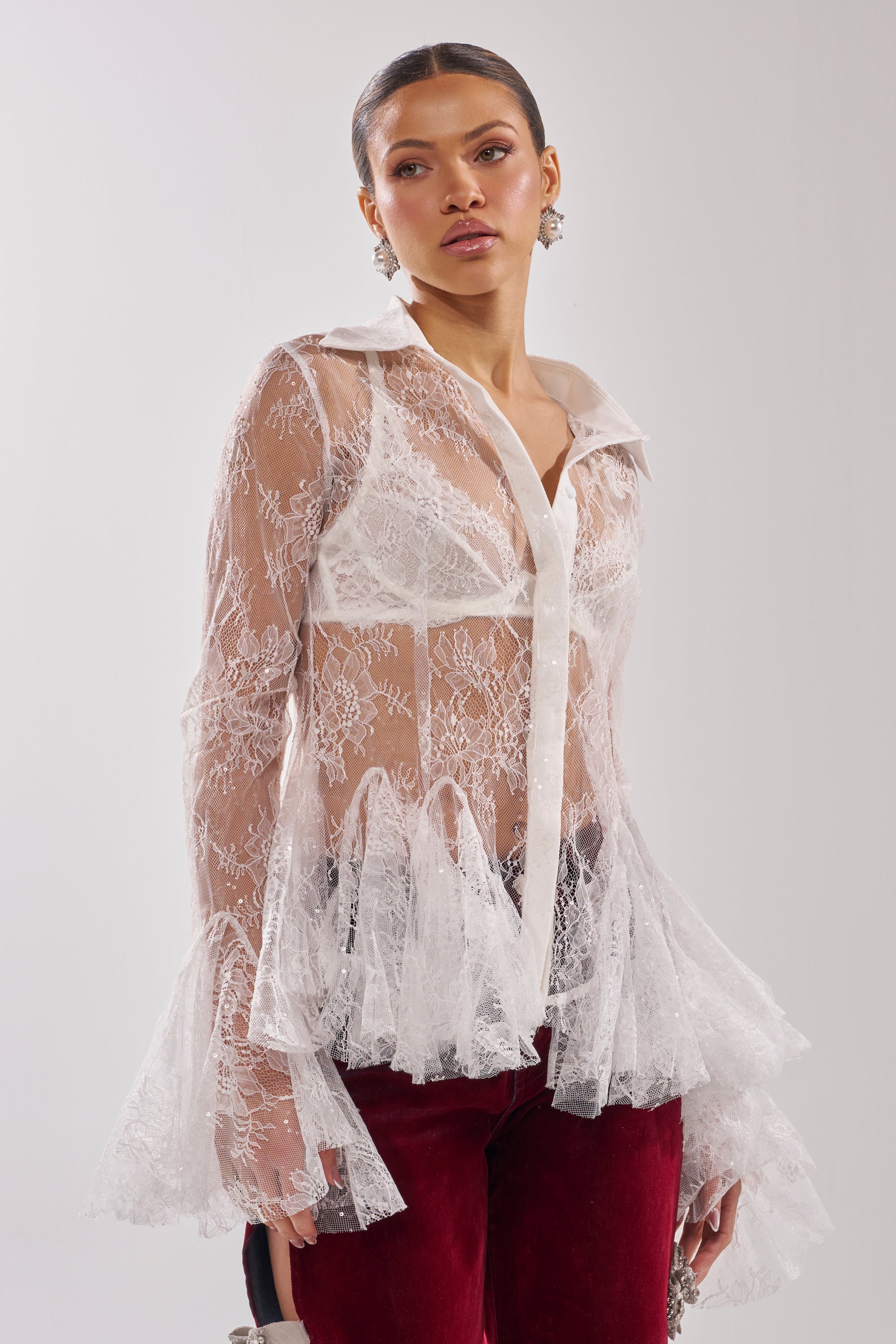 A woman models the LADY LOVER LACE BUTTON DOWN—a sheer lace blouse with flared cuffs and sequin accents—over a white bra, paired with red velvet pants, against a plain light background. 3
