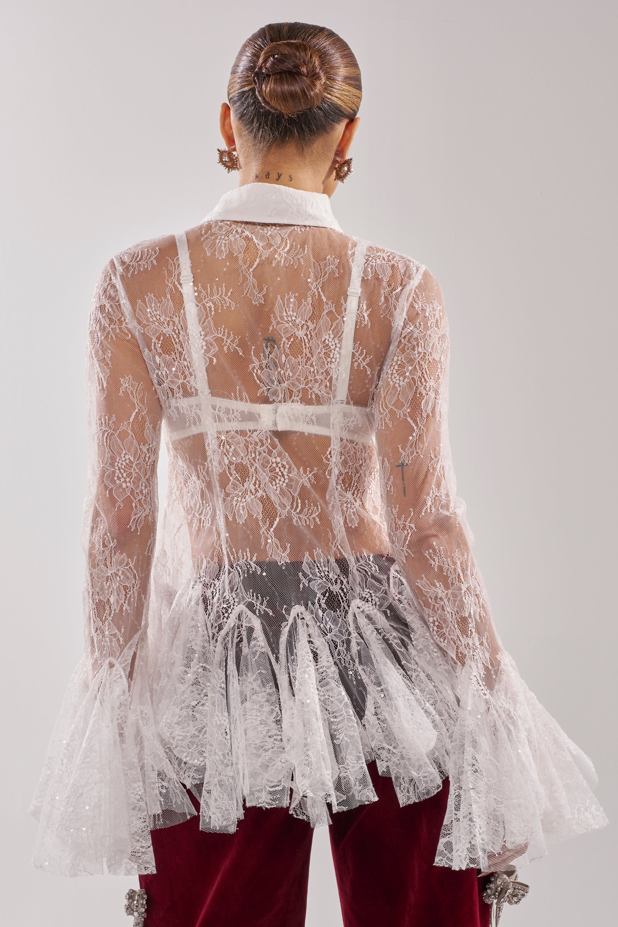 A person with hair in a sleek bun is seen from behind wearing the LADY LOVER LACE BUTTON DOWN over a white bra, featuring flared sleeves, paired with dark red velvet pants against a plain light gray background. 6