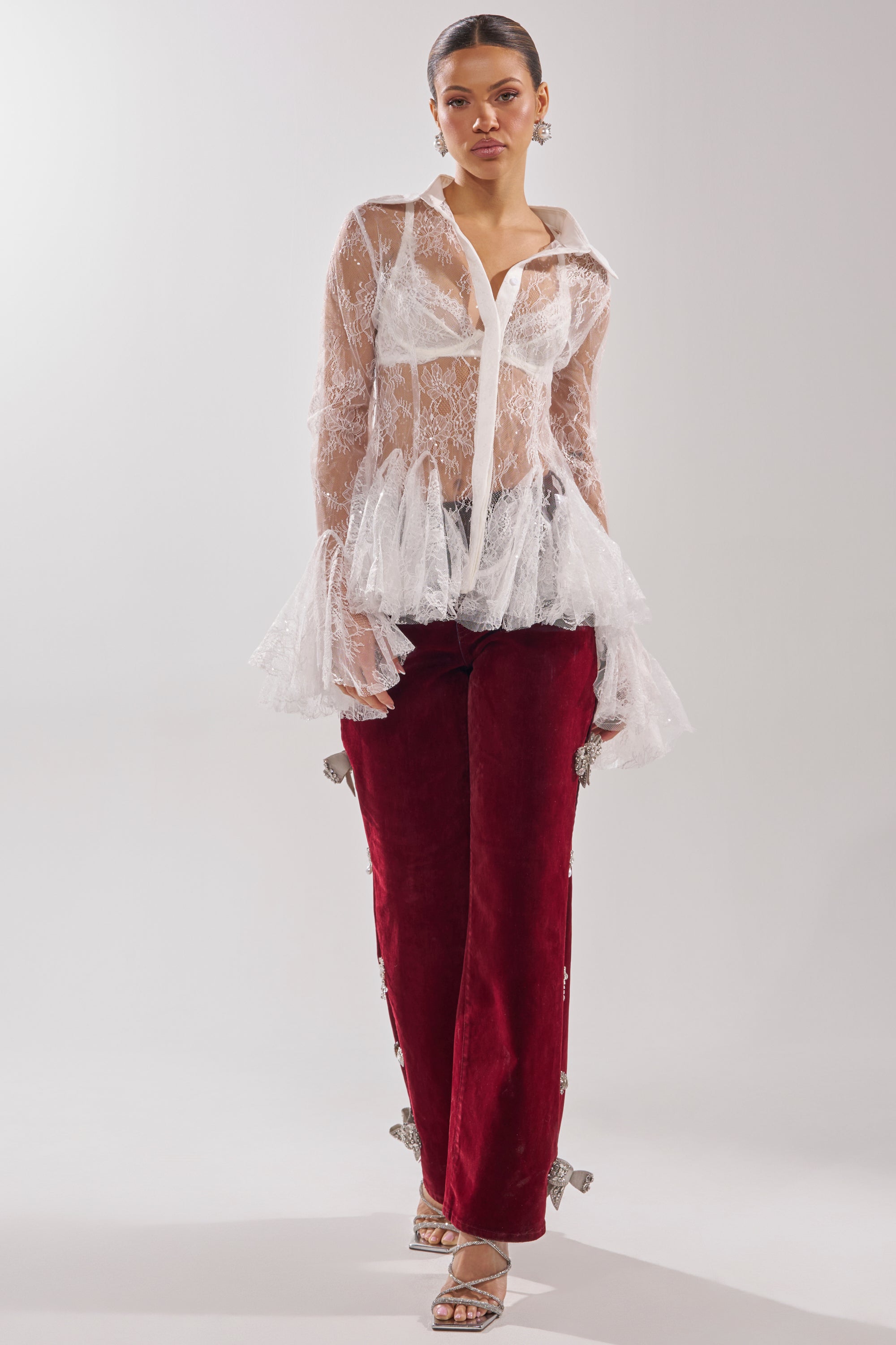 A woman models the LADY LOVER LACE BUTTON DOWN, a sheer lace blouse with flared cuffs, styled with burgundy velvet pants featuring decorative accents and strappy high-heeled sandals. 7