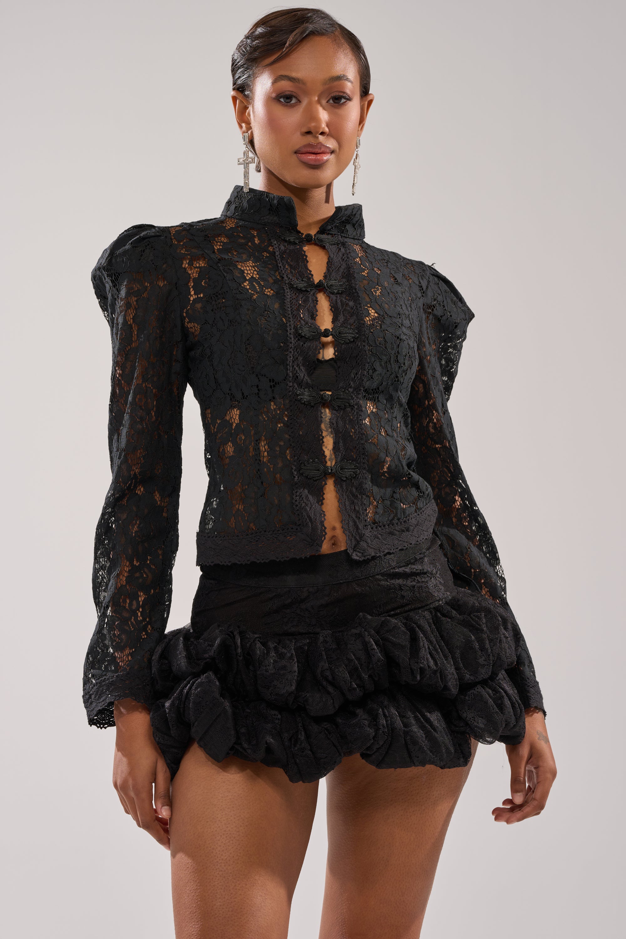 A woman confidently poses against a plain light background wearing the SEXY AS CAN BE LACE BLOUSE with puffed shoulders and a short, ruffled black skirt. 1