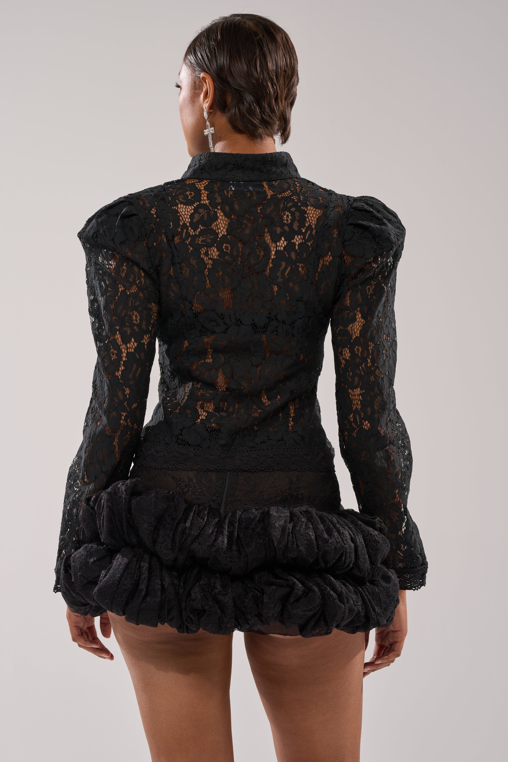 A person stands with their back to the camera, wearing the SEXY AS CAN BE LACE BLOUSE and a short black ruffled skirt against a plain, light-colored background. 3