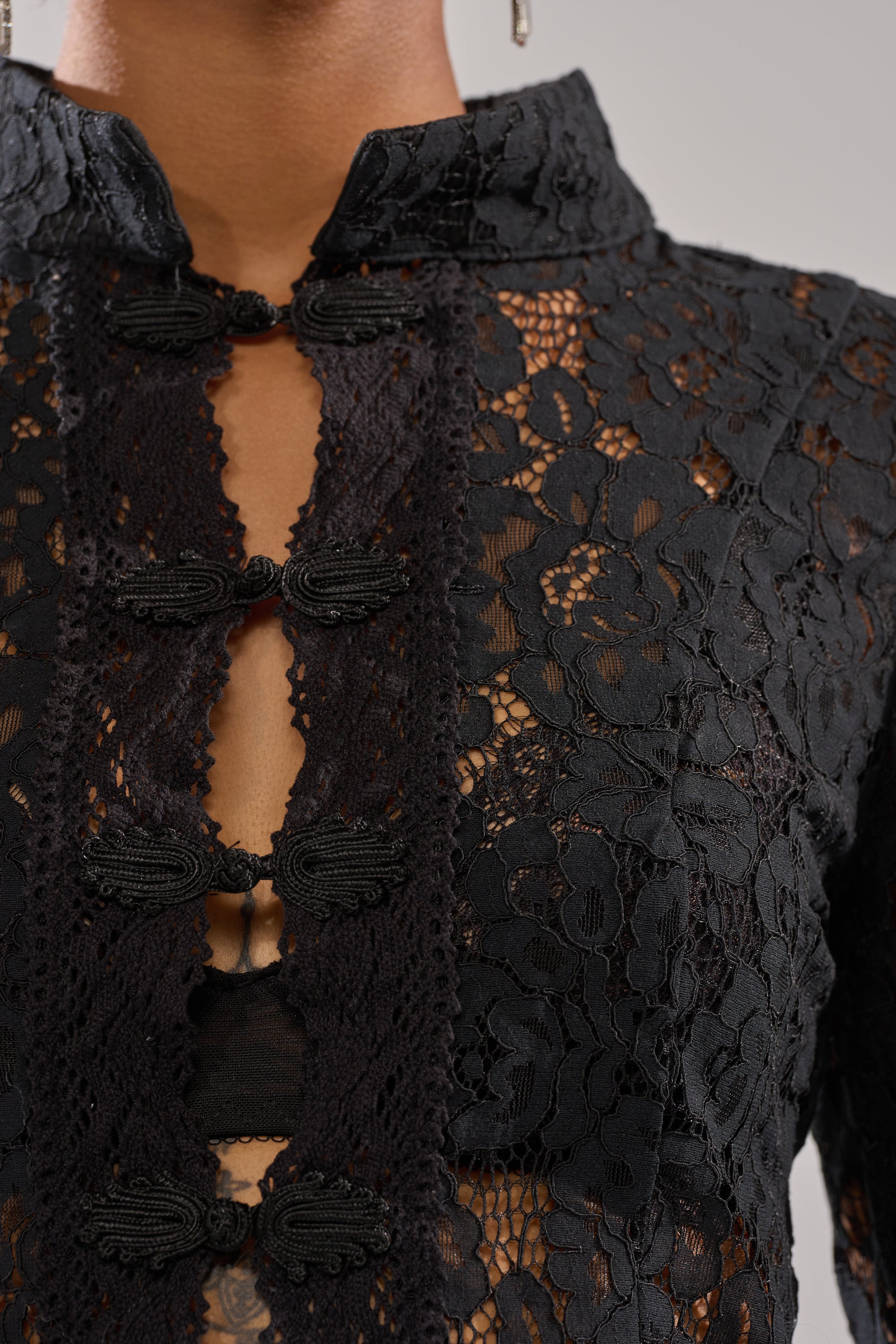 Close-up of a person wearing the SEXY AS CAN BE LACE BLOUSE, featuring black sheer floral lace, decorative looped buttons, and a high mandarin collar. The semi-transparent fabric subtly reveals skin beneath. 6