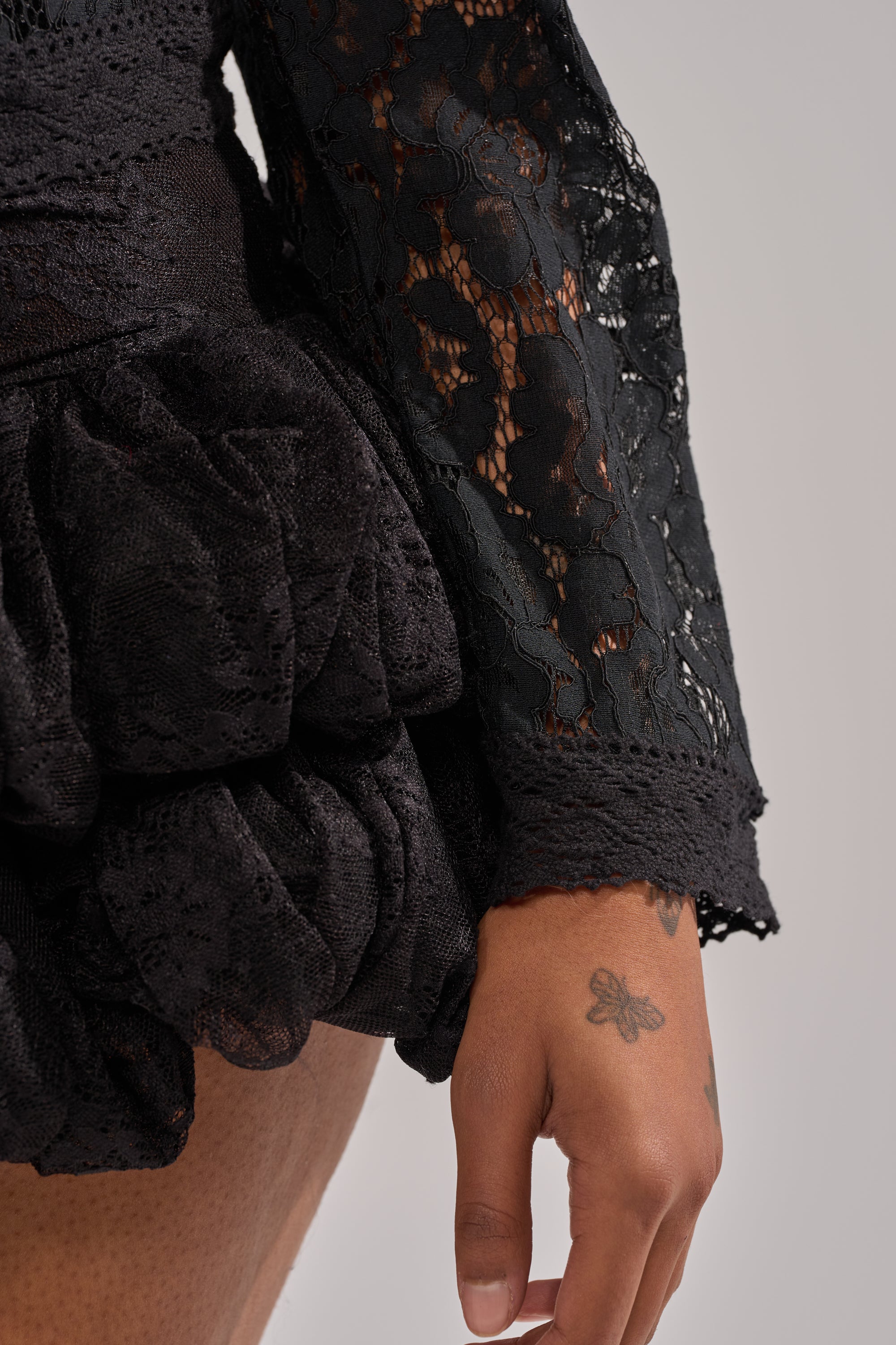A close-up of a person wearing the SEXY AS CAN BE LACE BLOUSE, featuring sheer floral lace and ruffled details, with their arm and hand—showing a small butterfly tattoo near the thumb—in view. 7