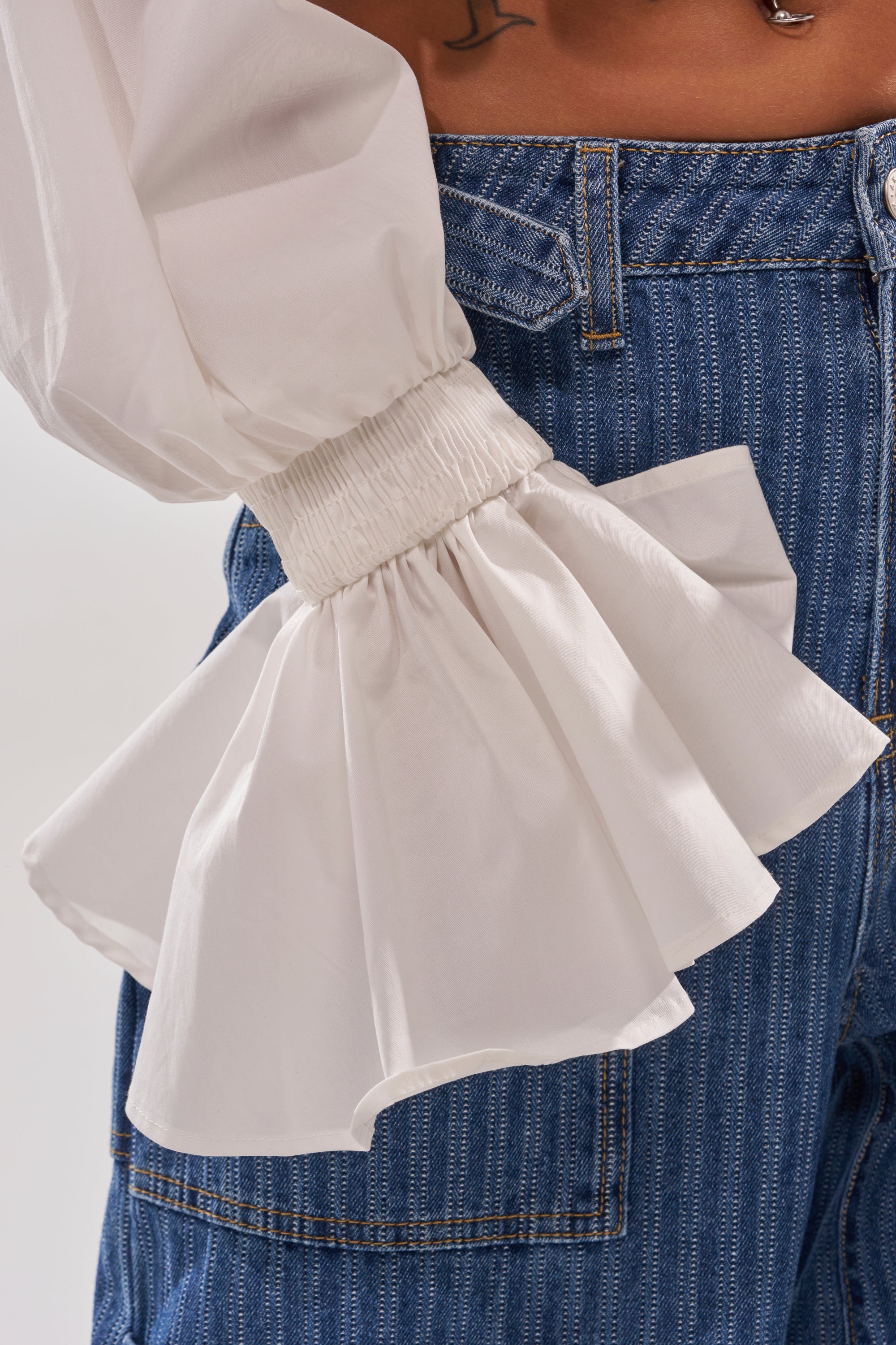 A close-up shows a person in blue jeans and the KELLY POPLIN BLOUSE, featuring white balloon sleeves with ruffled, gathered cuffs. The midriff and a tattoo are also partly visible at the top of the image.