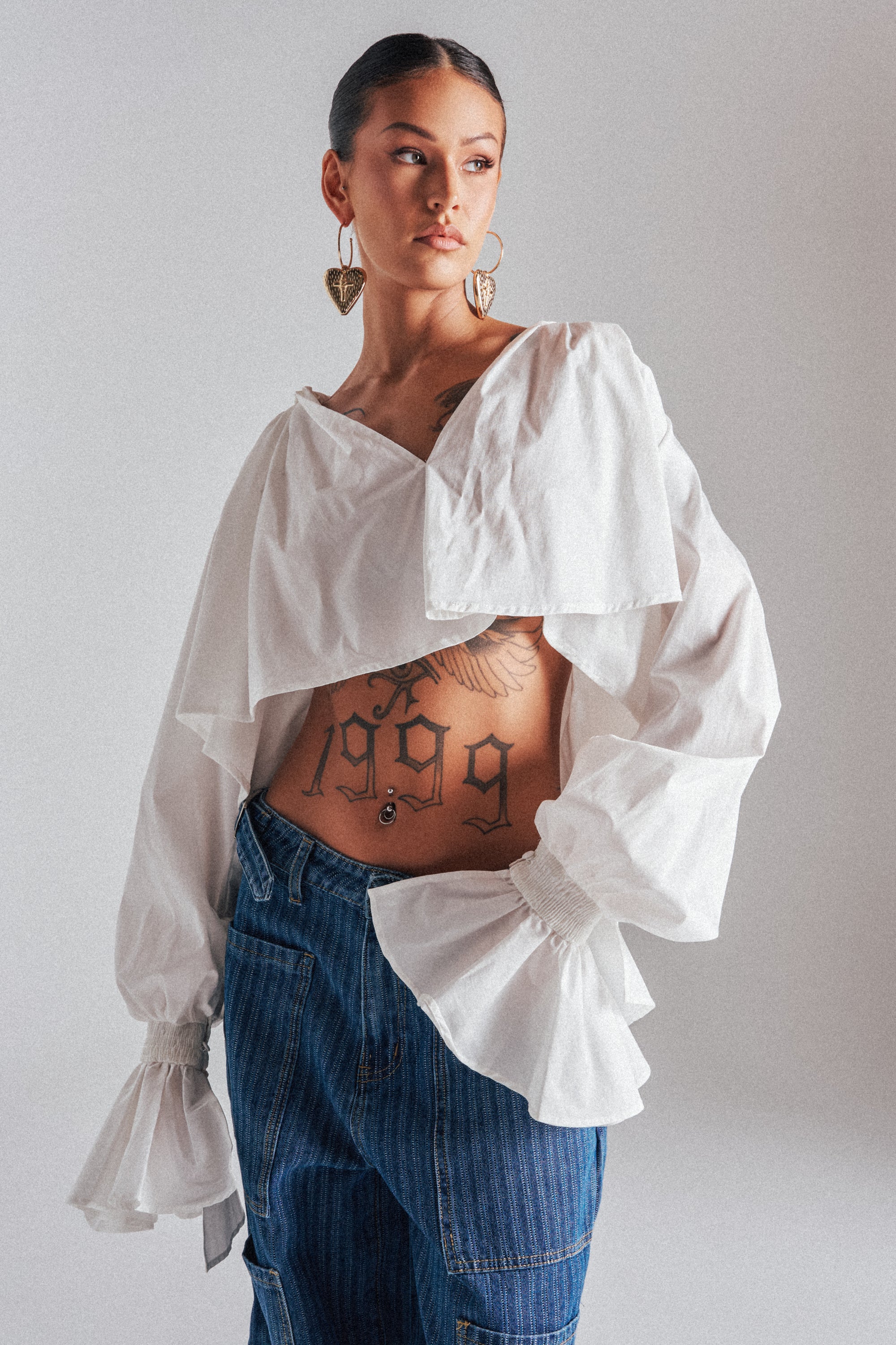 Wearing the KELLY POPLIN BLOUSE, a person with large gold heart-shaped earrings and blue jeans stands confidently, displaying a tattooed torso with "1999" against a plain background.