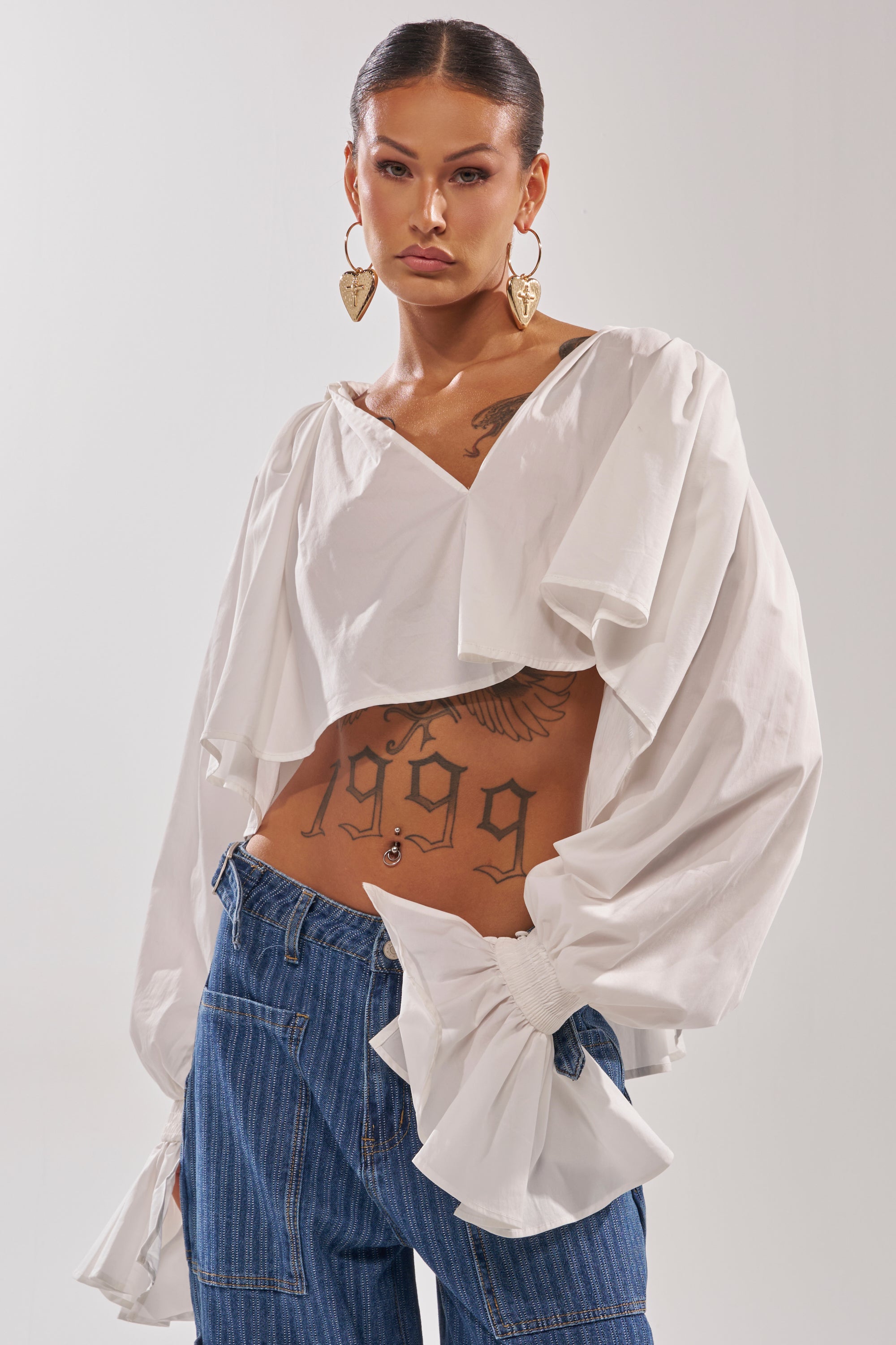 A woman with long dark hair in a sleek ponytail wears the KELLY POPLIN BLOUSE, a white cropped top with balloon sleeves, paired with blue jeans and large gold heart-shaped earrings; tattoos are visible on her chest and stomach.