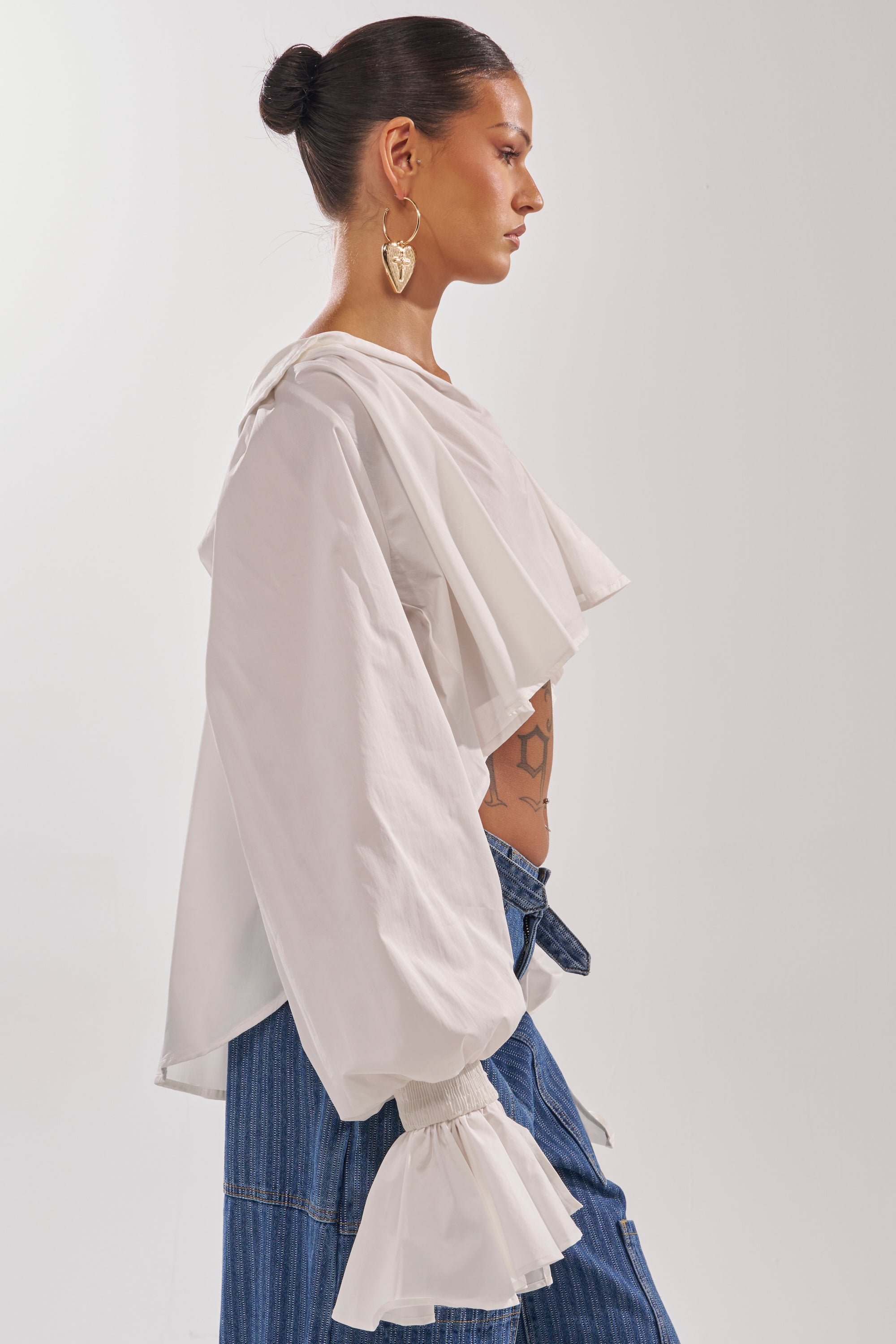 A woman in profile wears the KELLY POPLIN BLOUSE with balloon sleeves and ruffled cuffs, blue denim pants, gold heart-shaped earrings, and her hair in a neat bun against a plain background.