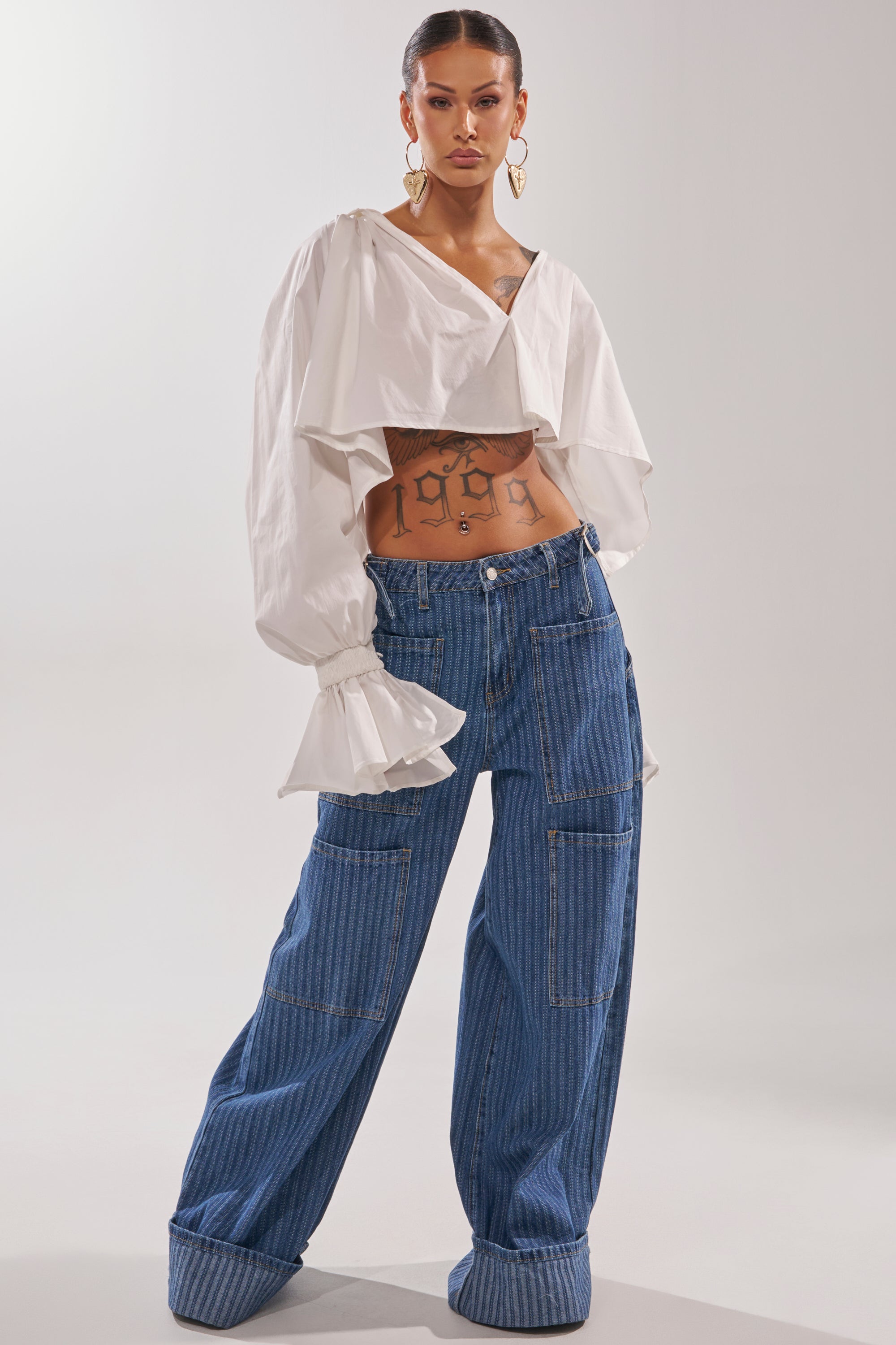 A woman poses confidently in the KELLY POPLIN BLOUSE, a white cropped style with ruffled long sleeves, paired with wide-legged blue denim jeans featuring large pockets. She accessorizes with gold hoop earrings and reveals a midriff tattoo.