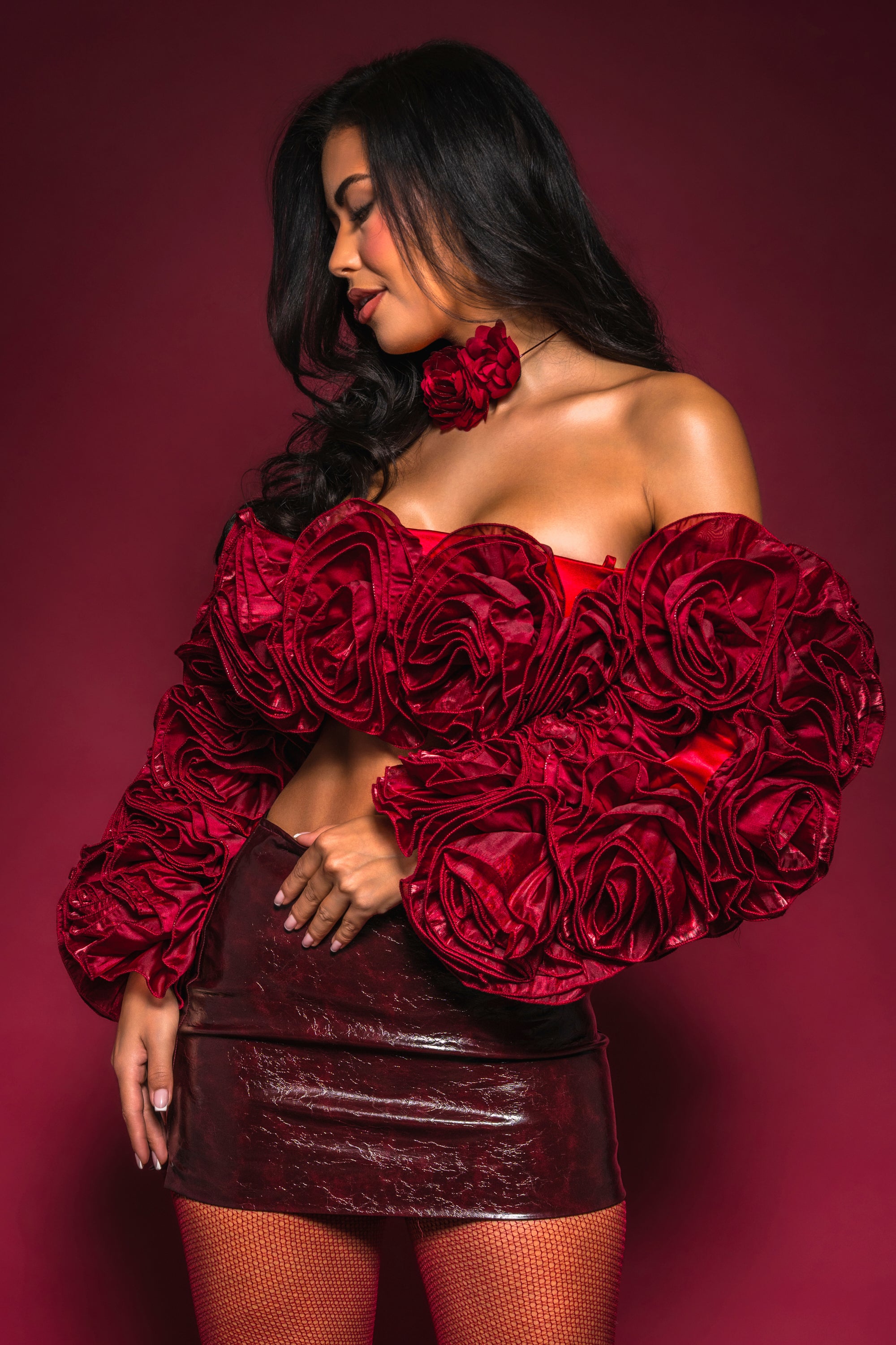 A woman poses against a maroon background, wearing the ROSES FOR EVERYONE BLOUSE with 3D rosette details, a red flower choker, shiny burgundy mini skirt, and fishnet stockings.